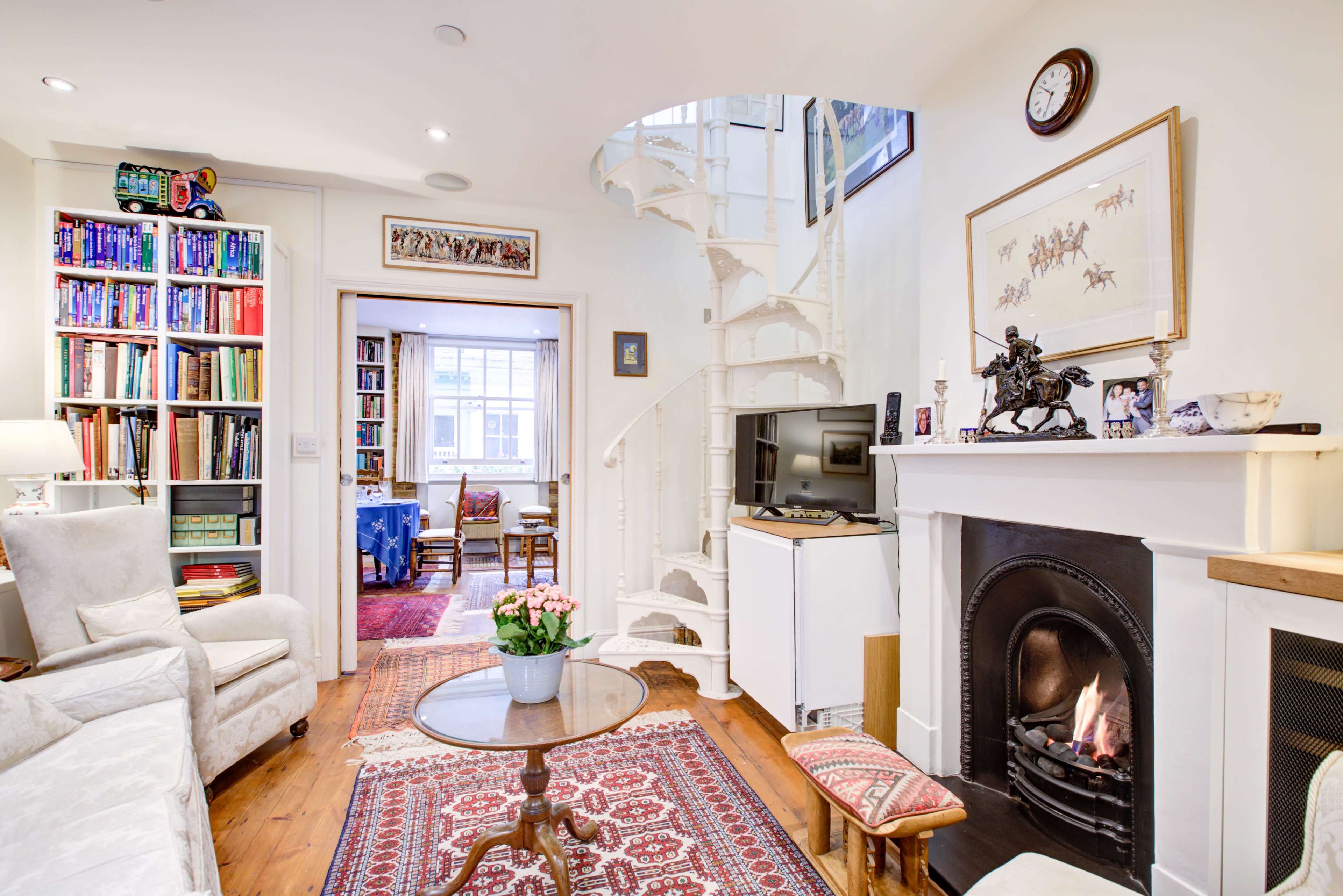 A cozy interior with a spiral staircase, a white bookshelf filled with books, a fireplace with a small flame, and a sitting area adorned with rugs and furniture.
