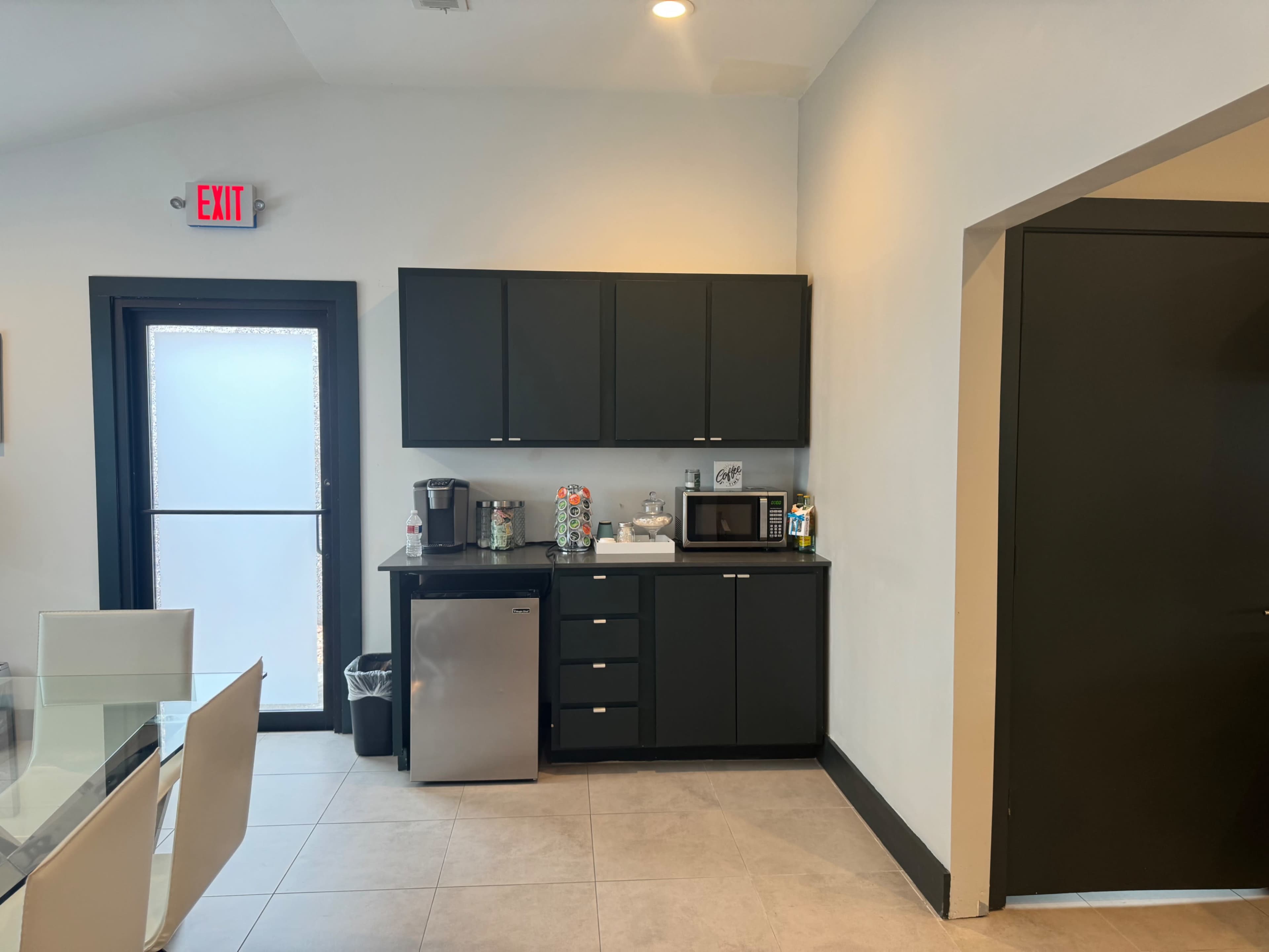 The image shows a modern kitchen area with black cabinetry, a silver refrigerator, a microwave, and a glass door leading outside.
