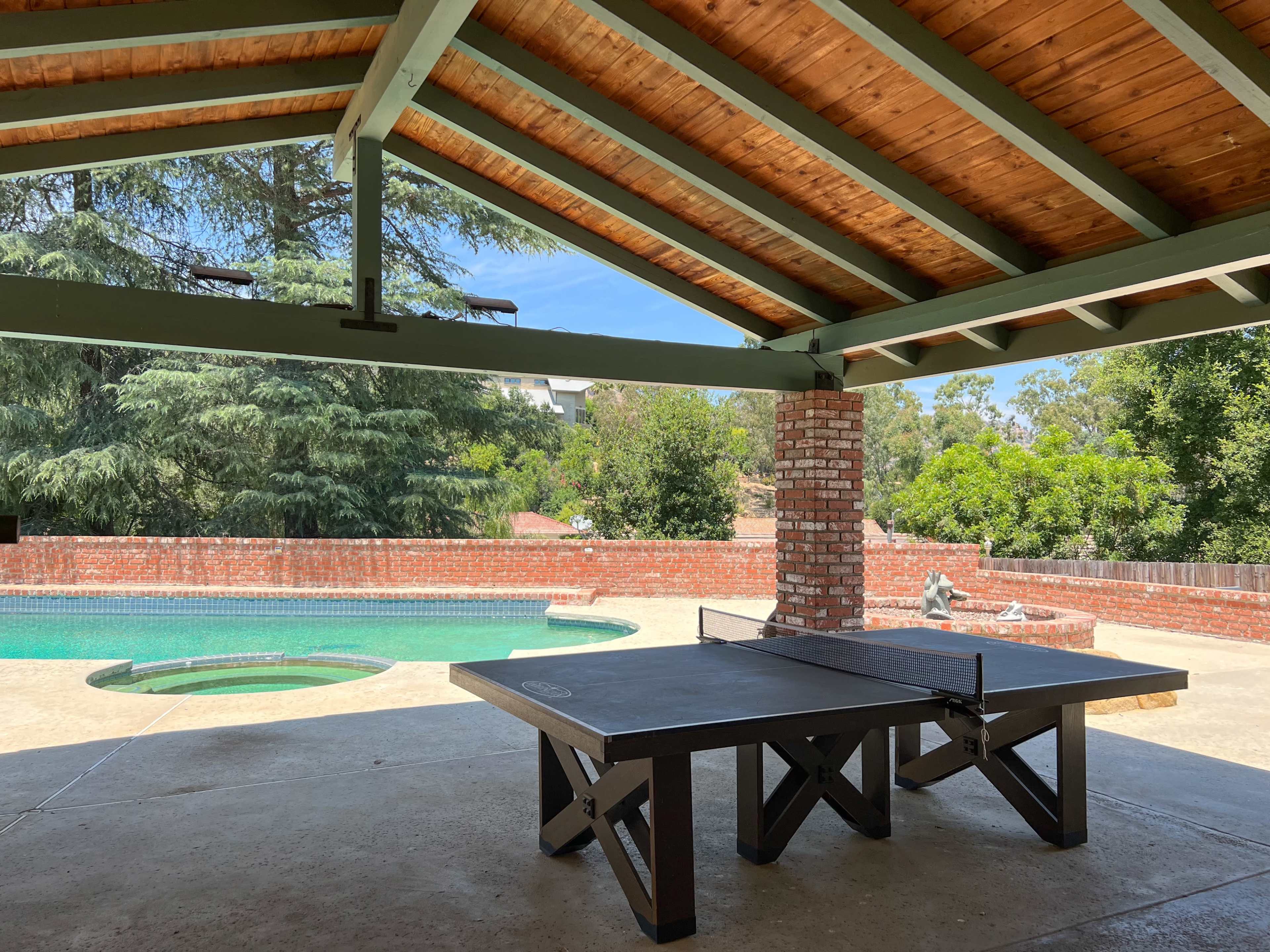 A shaded outdoor area features a ping pong table with a pool and landscaped greenery visible in the background.