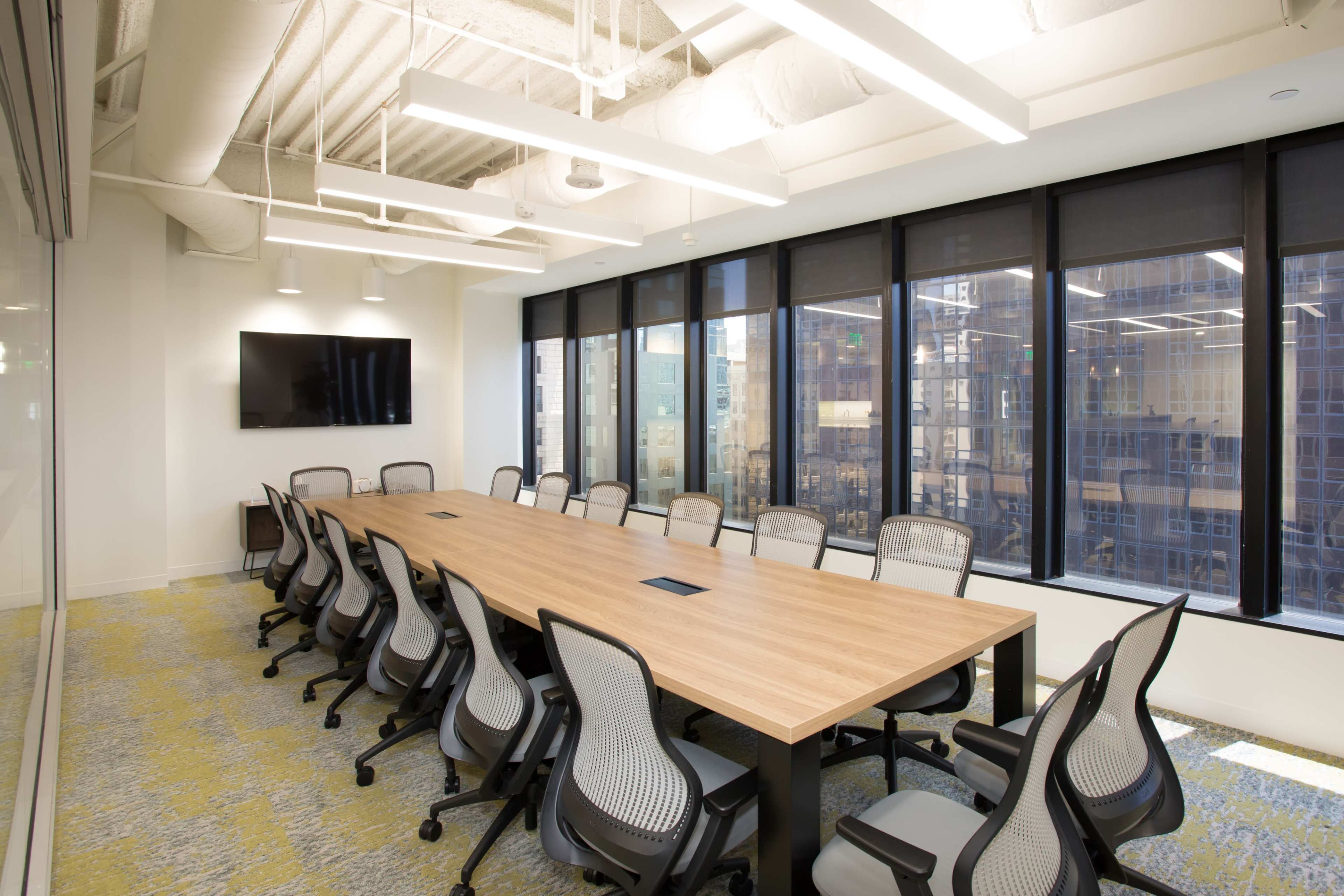 A large conference room features a long wooden table surrounded by ergonomic chairs and large windows with city views.