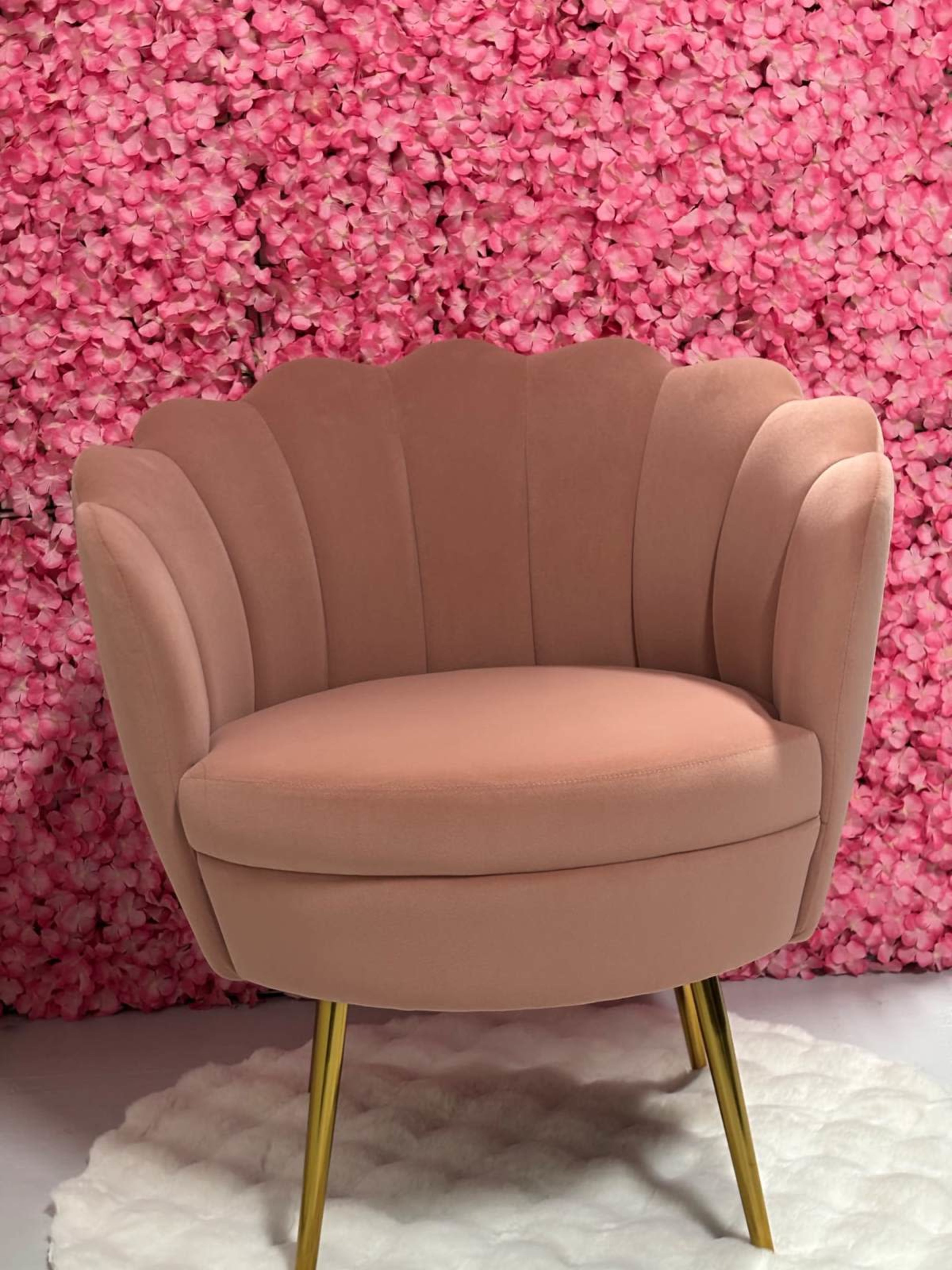 A pink fabric chair with a scalloped back sits on a white rug in front of a pink flower backdrop.