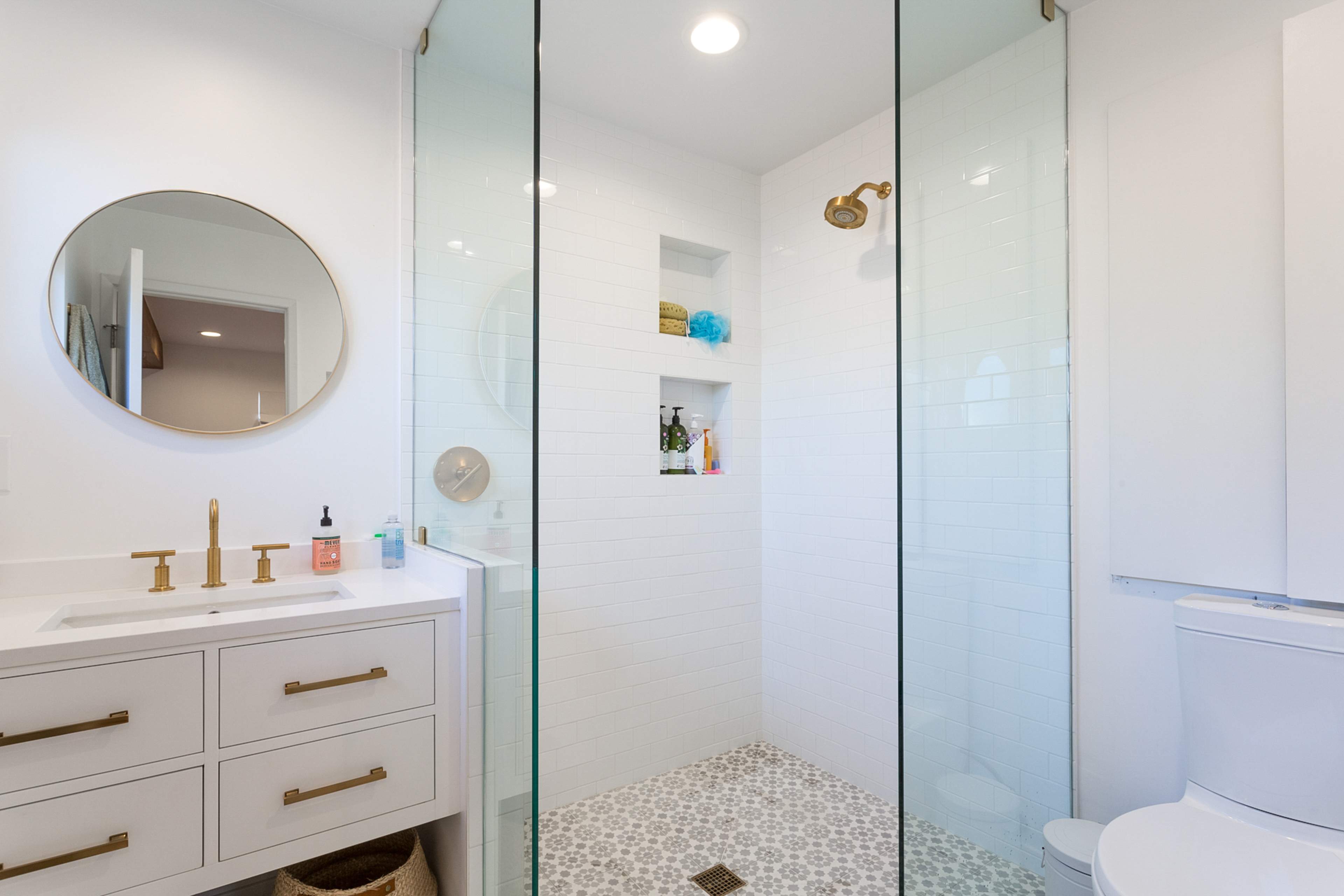 The bathroom features a glass-walled shower area, a round mirror above a white vanity with brass faucets, and light tile flooring.