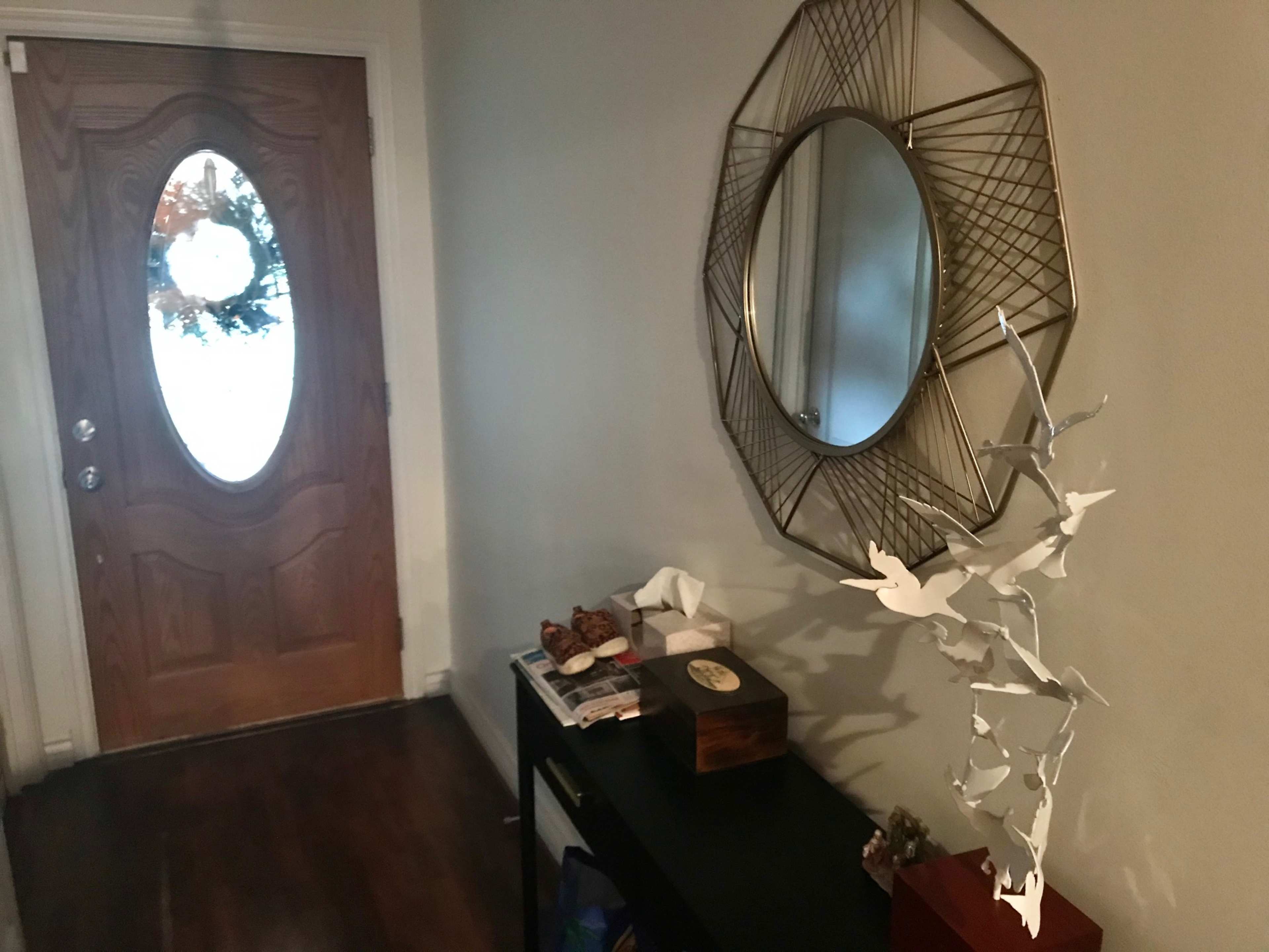 A hallway with a wooden front door, a decorative mirror on the wall, a small table with tissues and magazines, and a white bird sculpture.