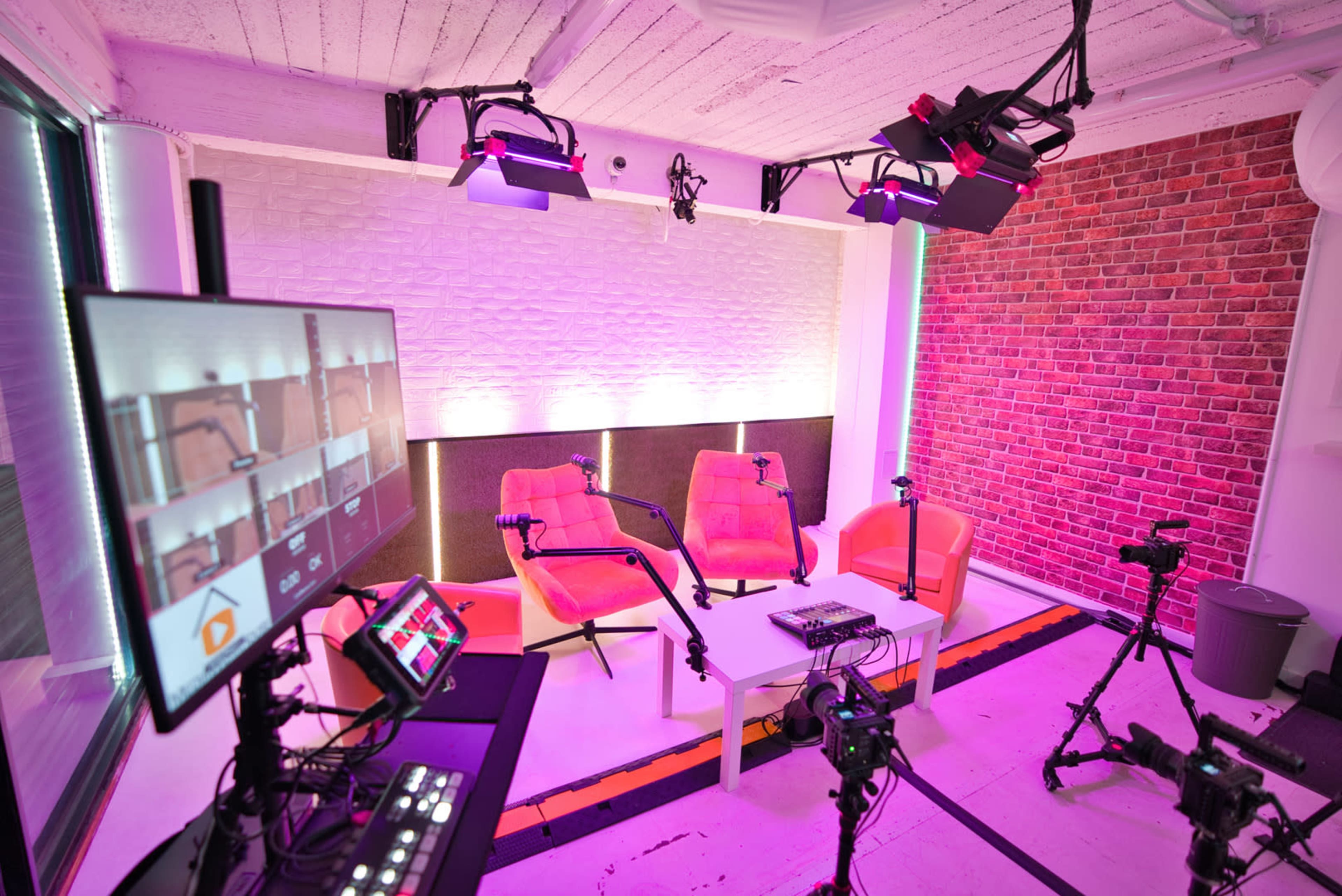 A modern podcast studio features two orange chairs, a white table, and multiple cameras and lighting equipment arranged around the space.