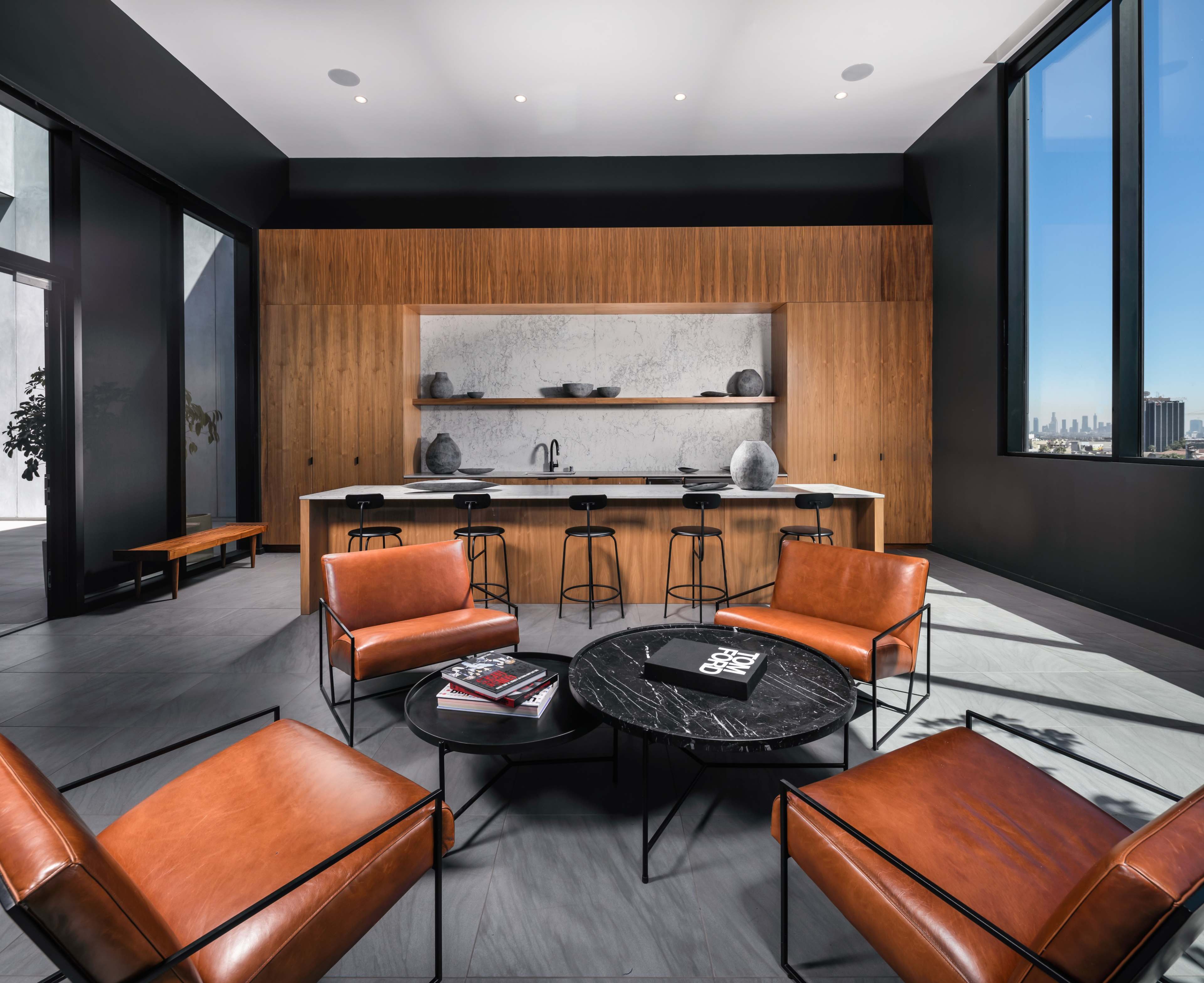 A modern living space features brown leather chairs and a sleek kitchen island with a marble countertop and tall windows offering a cityscape view.