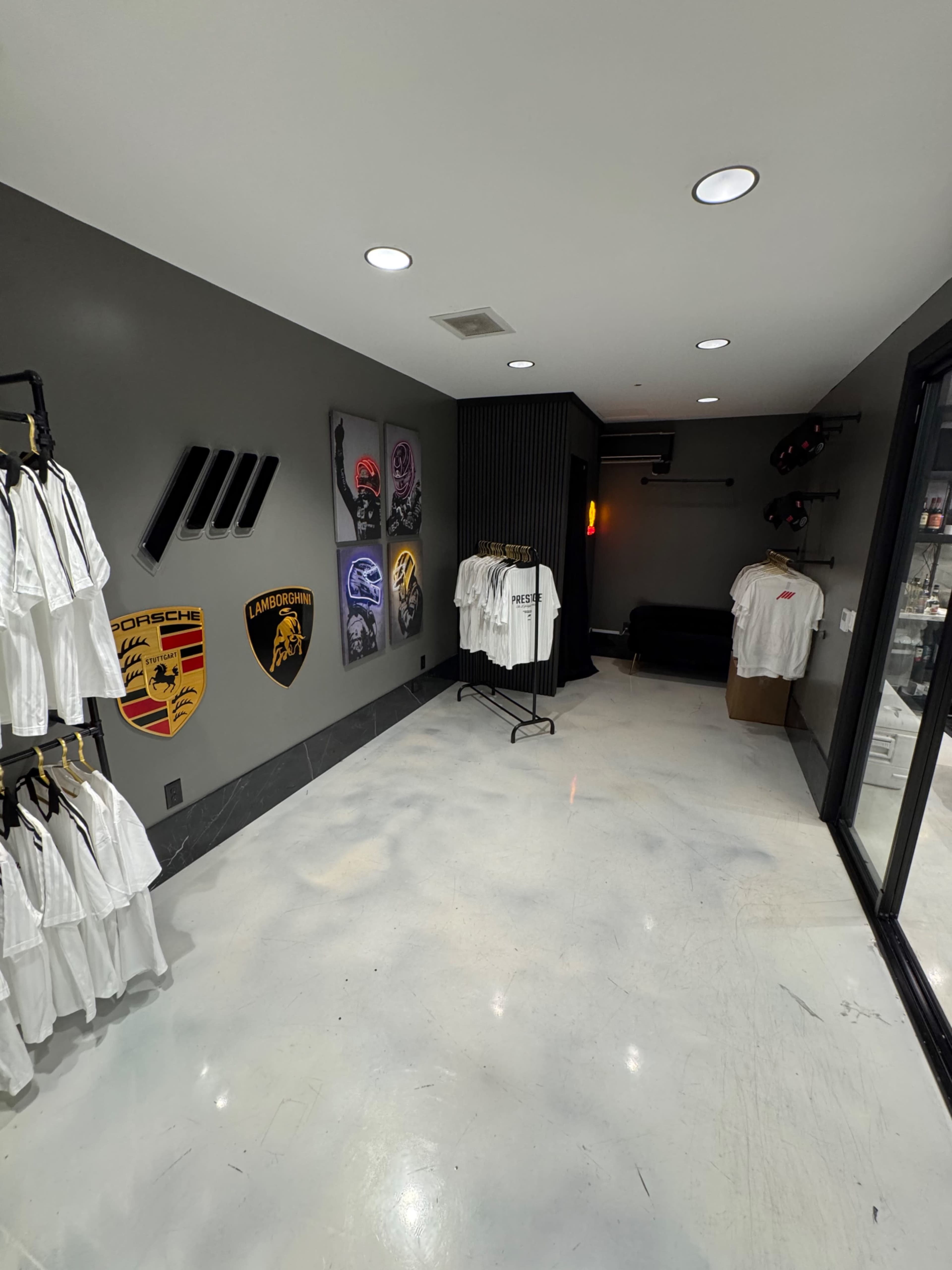 The interior of a retail space features a polished concrete floor, gray walls adorned with car brand logos, and racks displaying T-shirts.