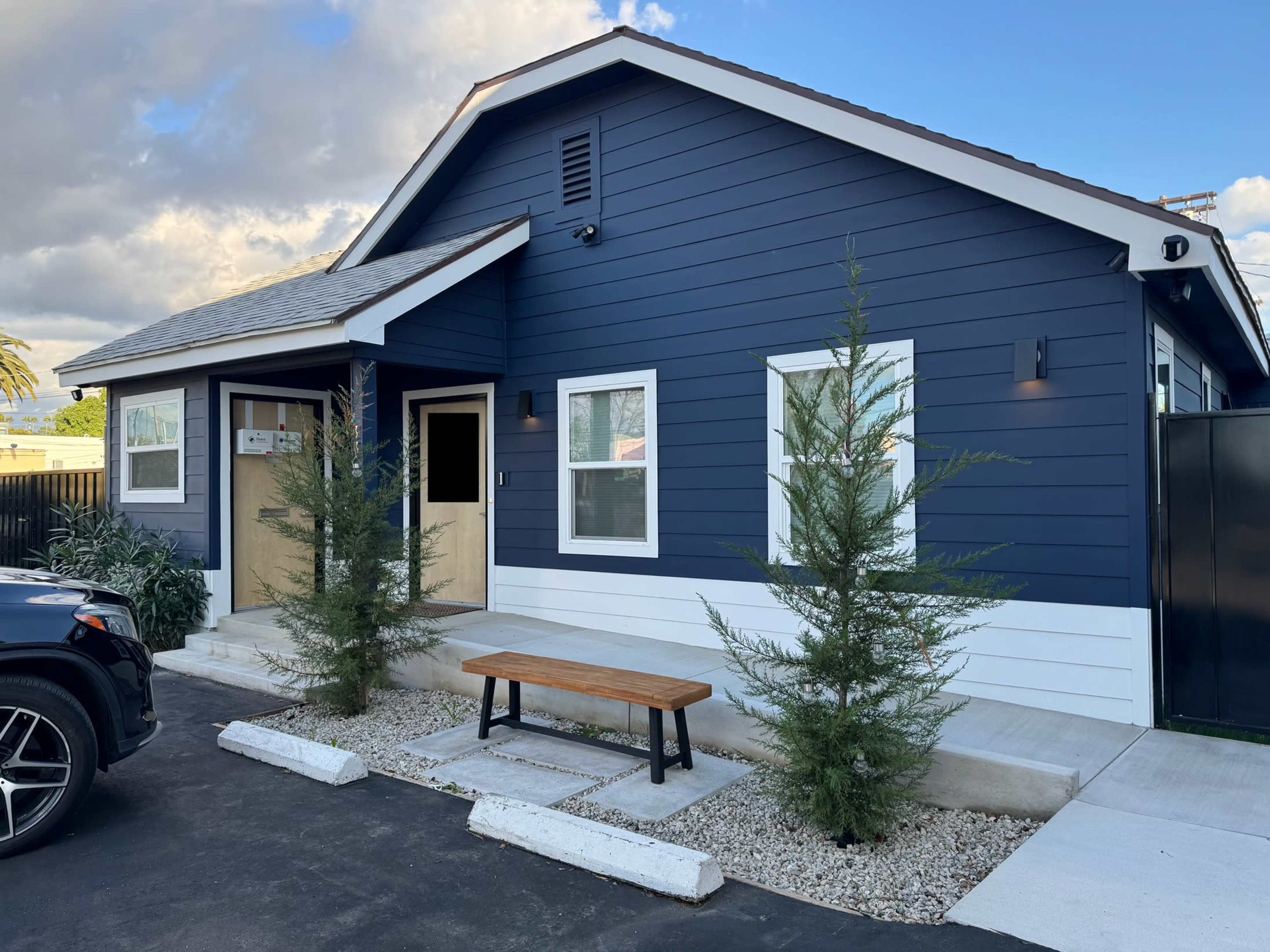 A blue and white house with two windows, a wooden bench in front, and landscaped trees beside the entrance.