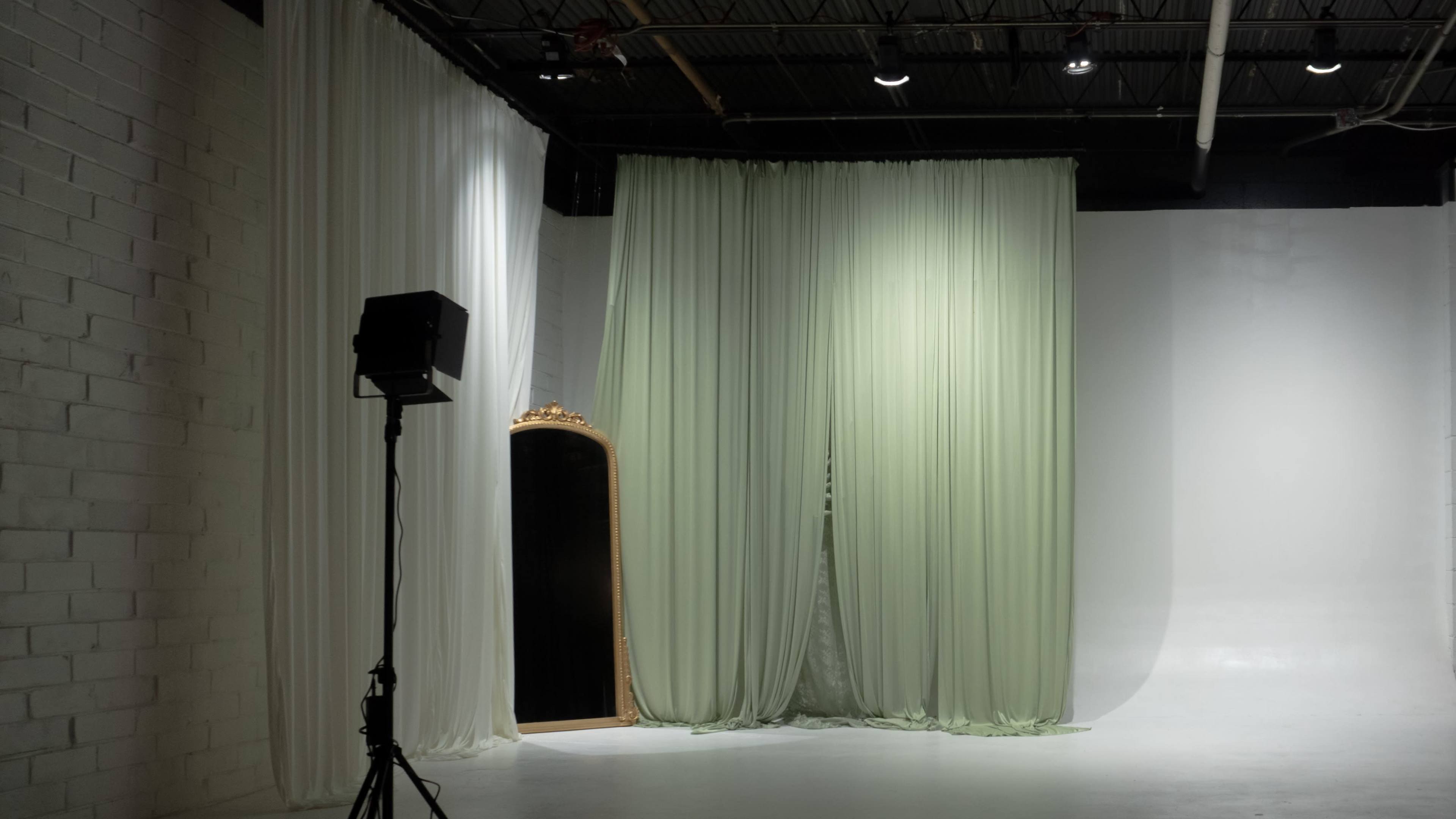 The image shows a spacious studio with white walls, large fabric drapes in light green and white, and a spotlight positioned on a stand.