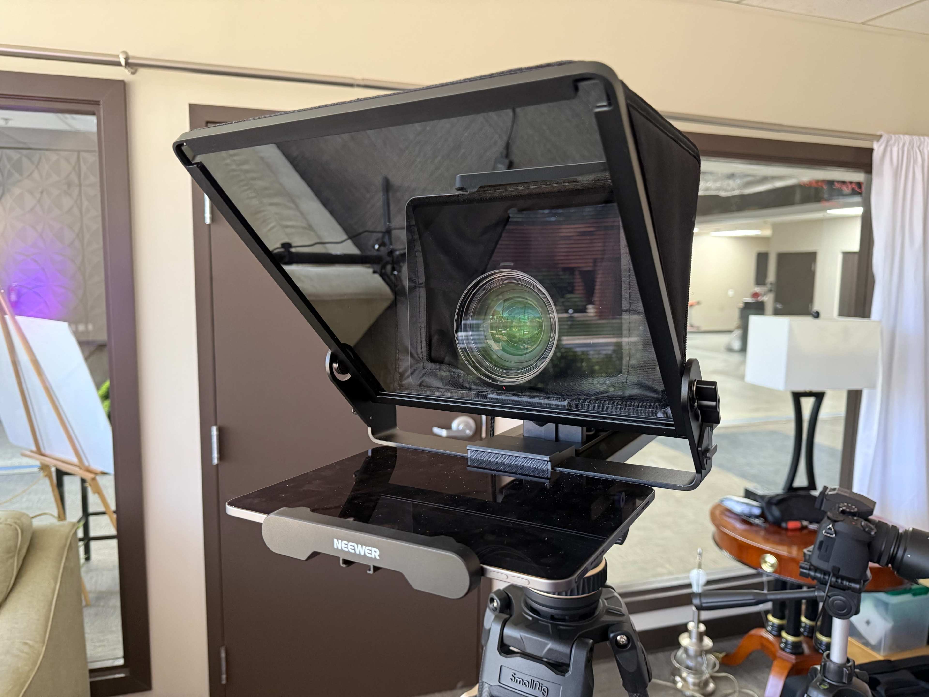 A teleprompter is set up on a tripod, displaying text and reflecting light from a camera lens inside a modern indoor space.