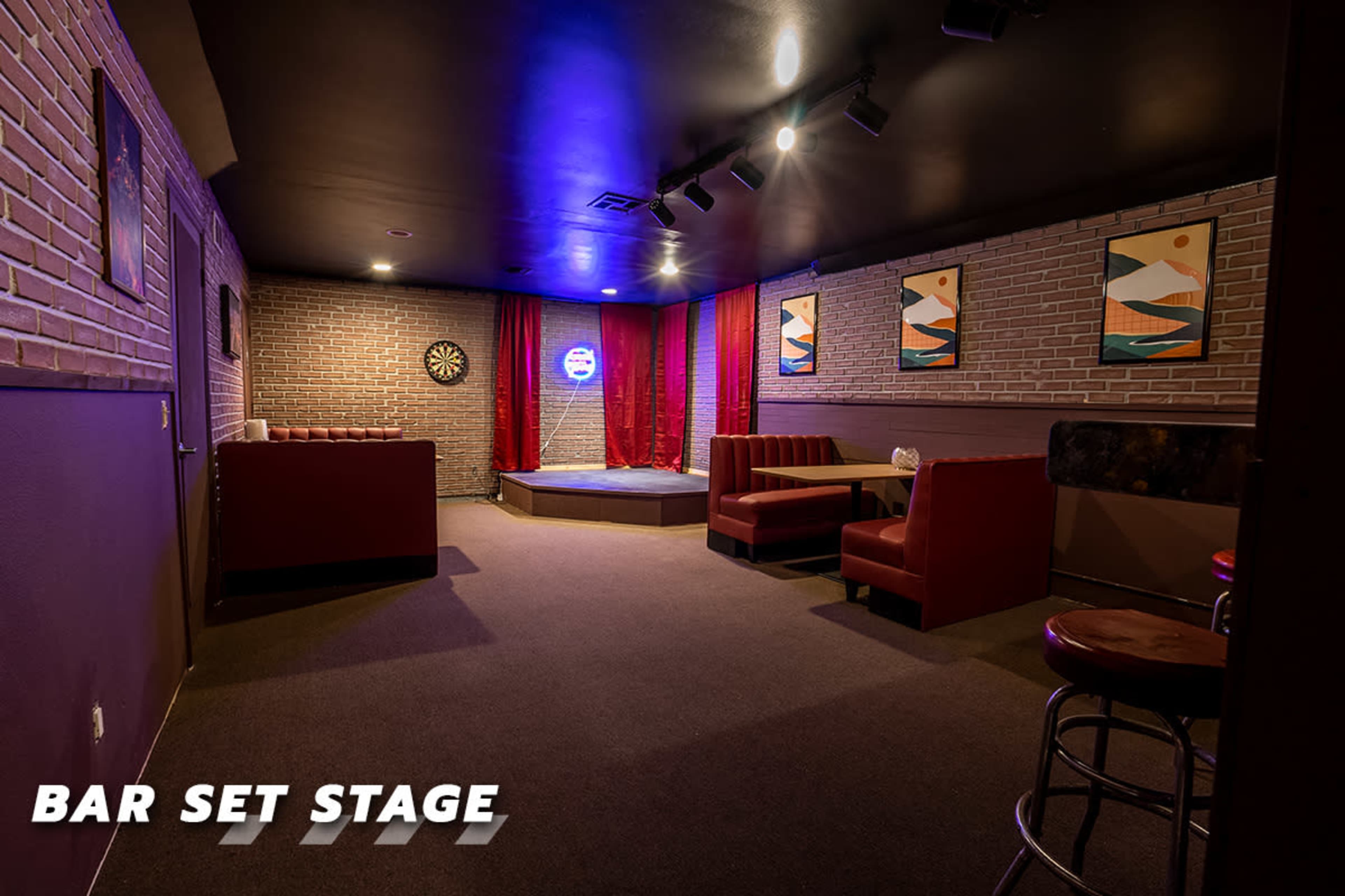 The image shows a small bar set stage with red seating, a raised performance area, and framed artwork on the walls.