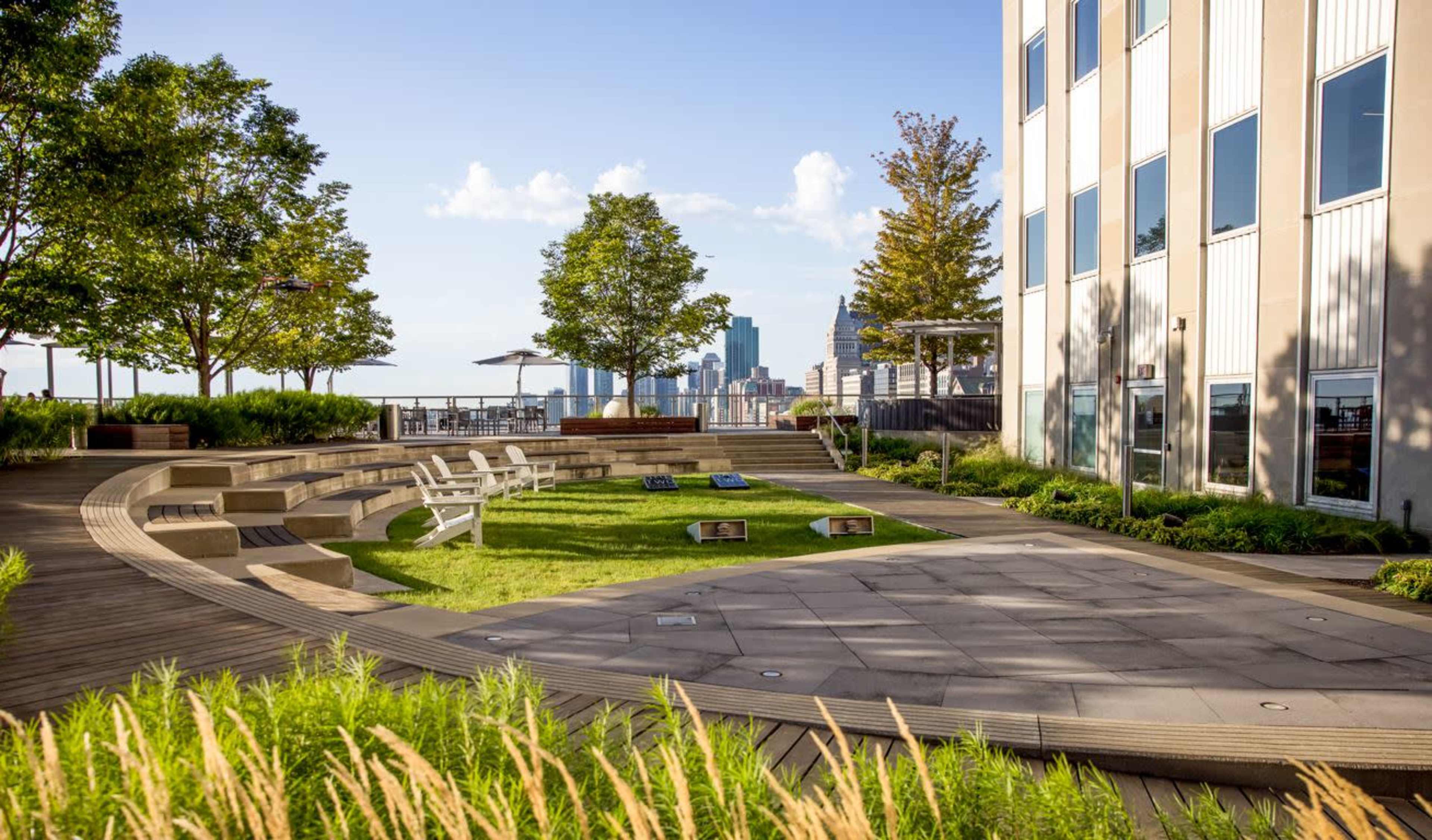 A landscaped rooftop garden with seating areas, greenery, and a city skyline in the background.