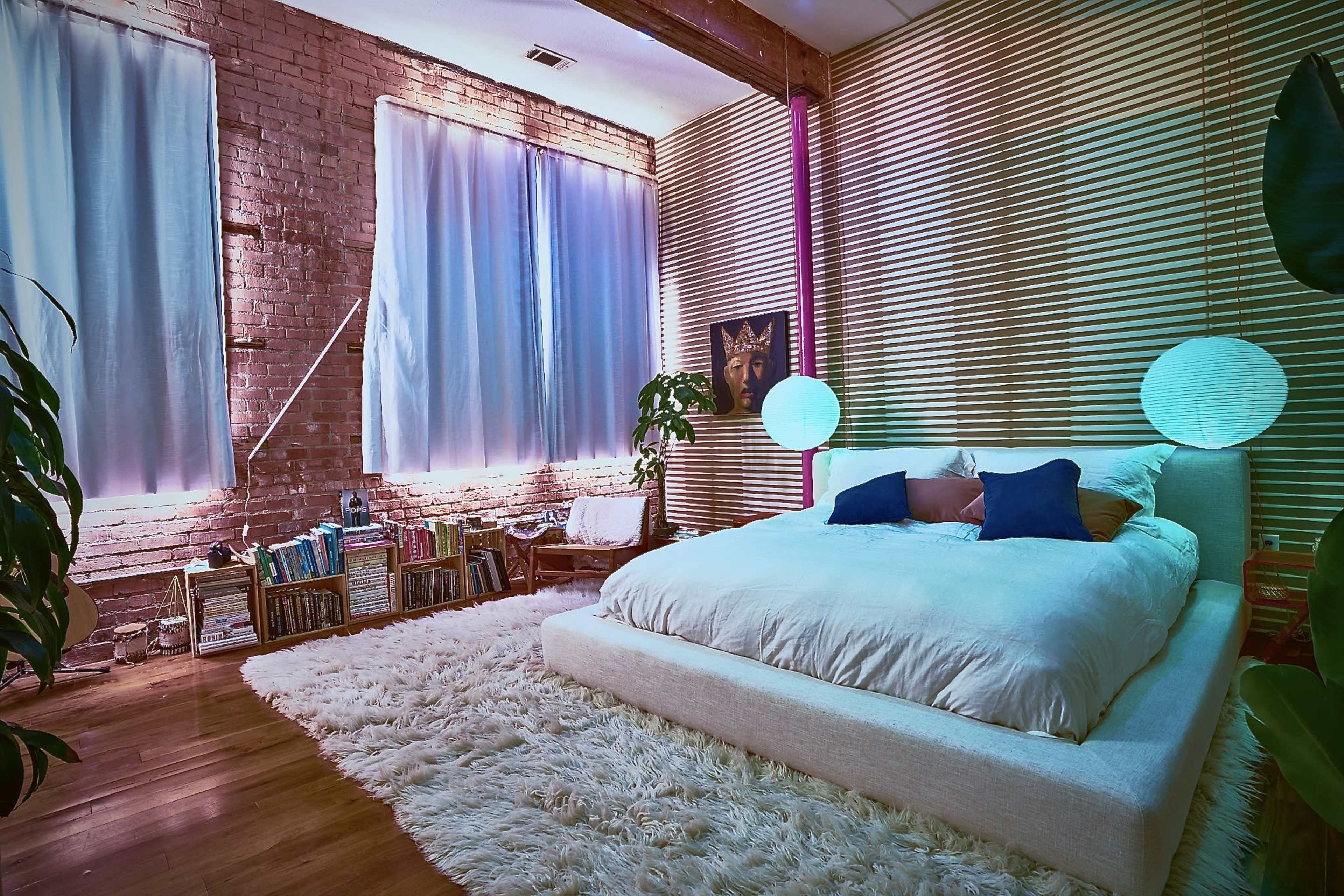 A cozy bedroom featuring a large bed with white bedding, a shaggy rug on the wooden floor, and shelves filled with books against a textured brick wall.