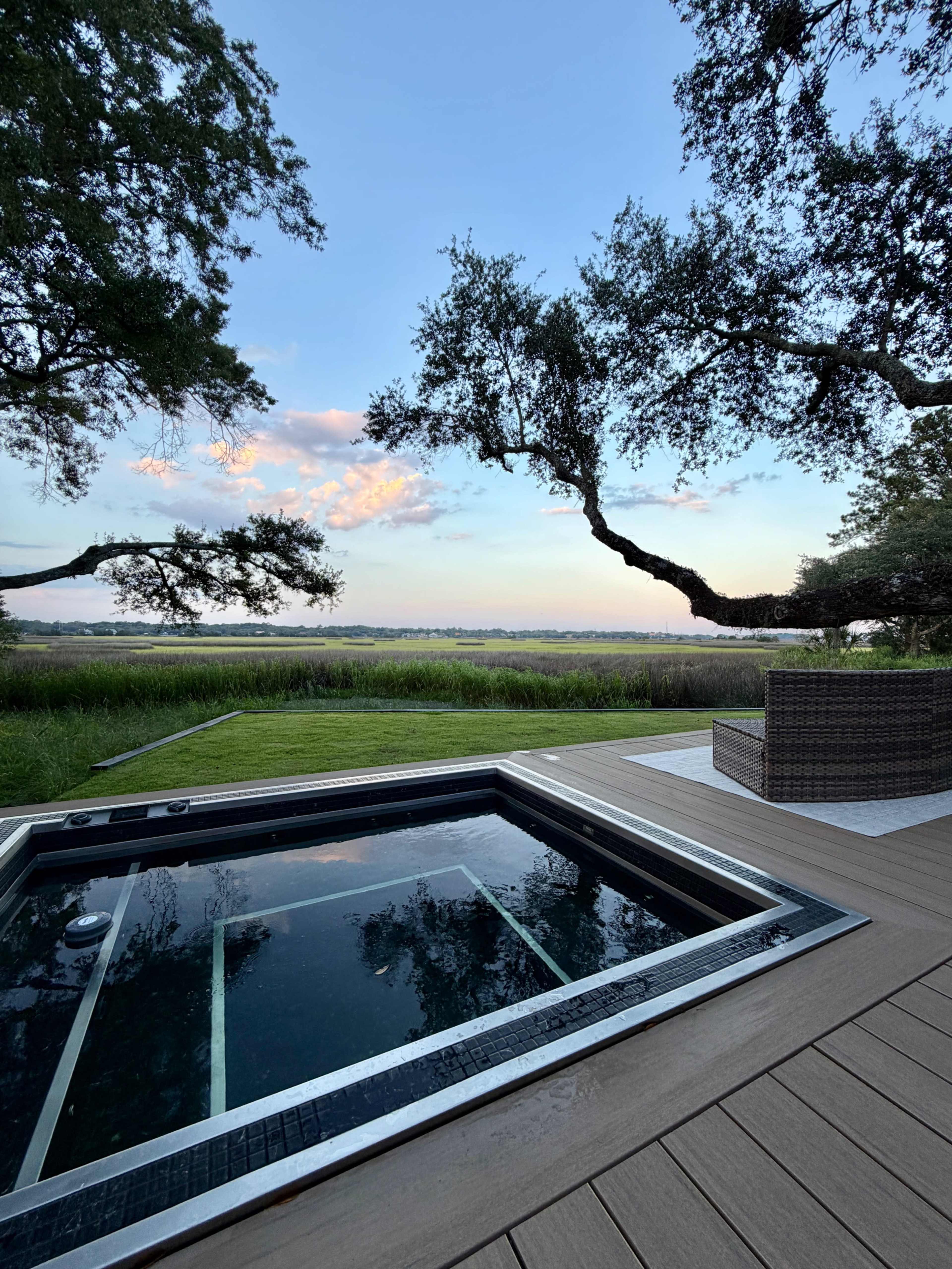 A hot tub sits on a wooden deck overlooking a grassy area and a calm body of water under a blue sky with scattered clouds.