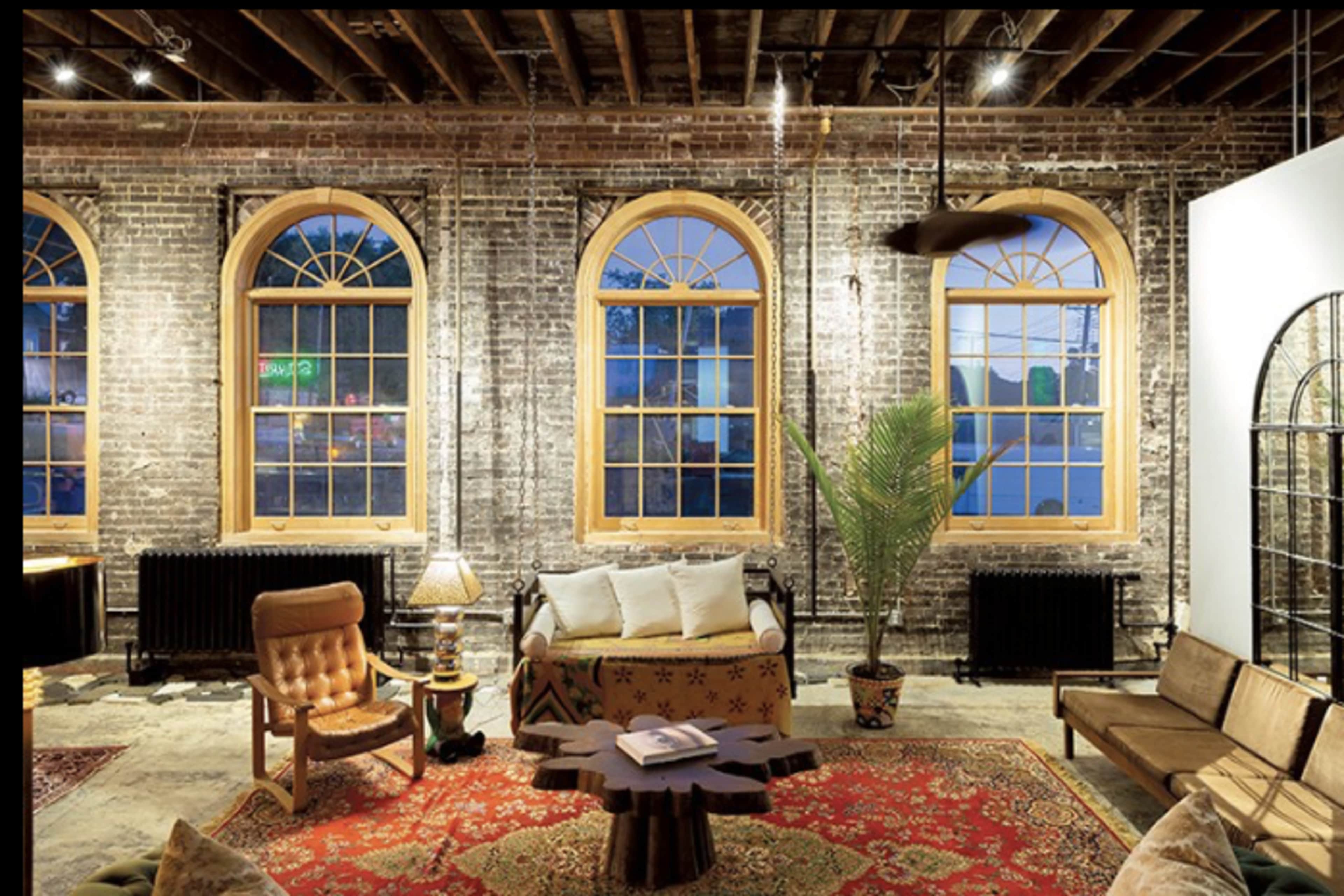 The image depicts a stylish living space with large, arched windows, exposed brick walls, and various seating arrangements on a patterned rug.