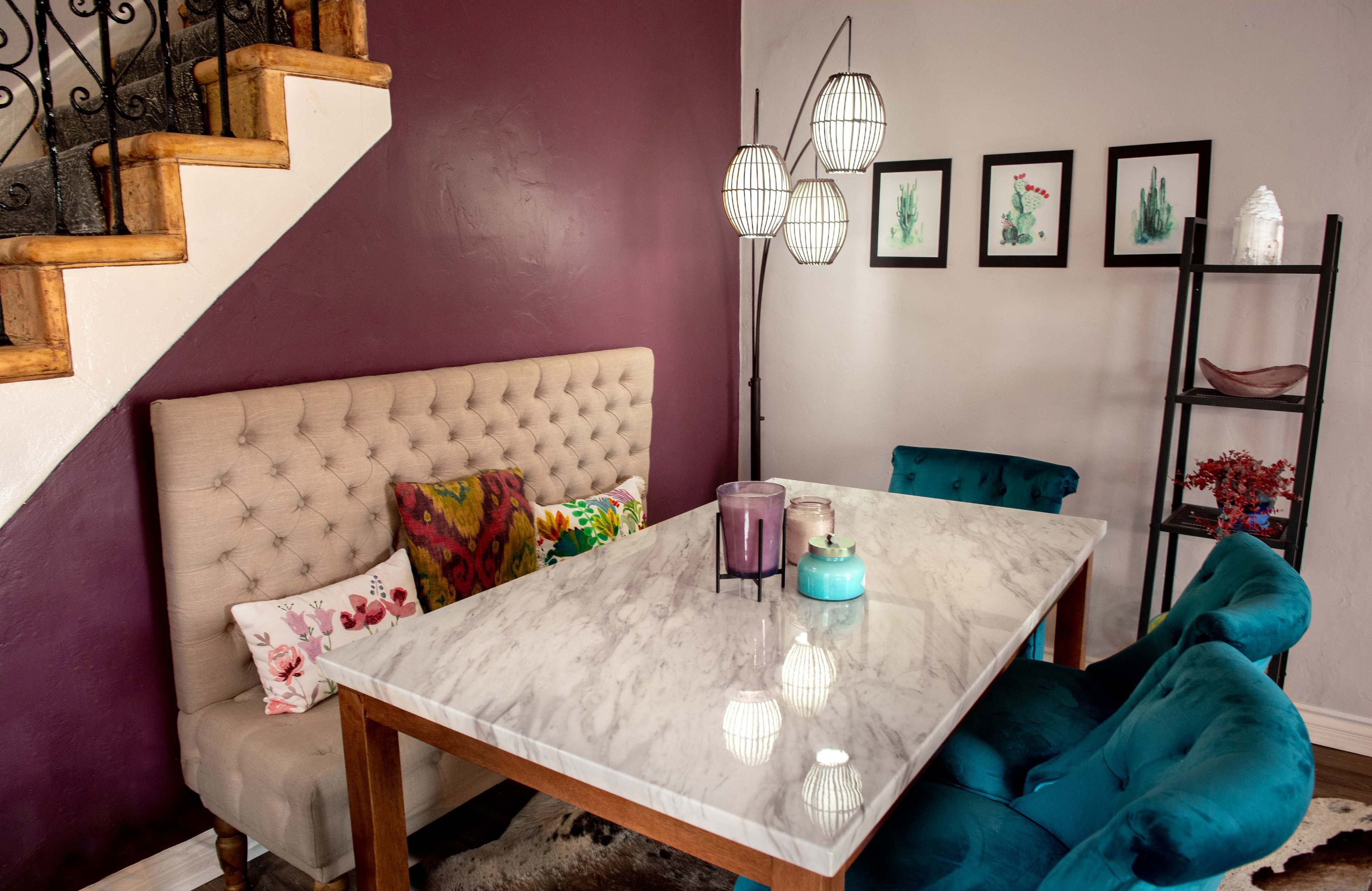A dining area features a marble-top table surrounded by teal chairs and a tufted bench against a deep purple wall, with decorative plants framed on the wall and a staircase in the background.