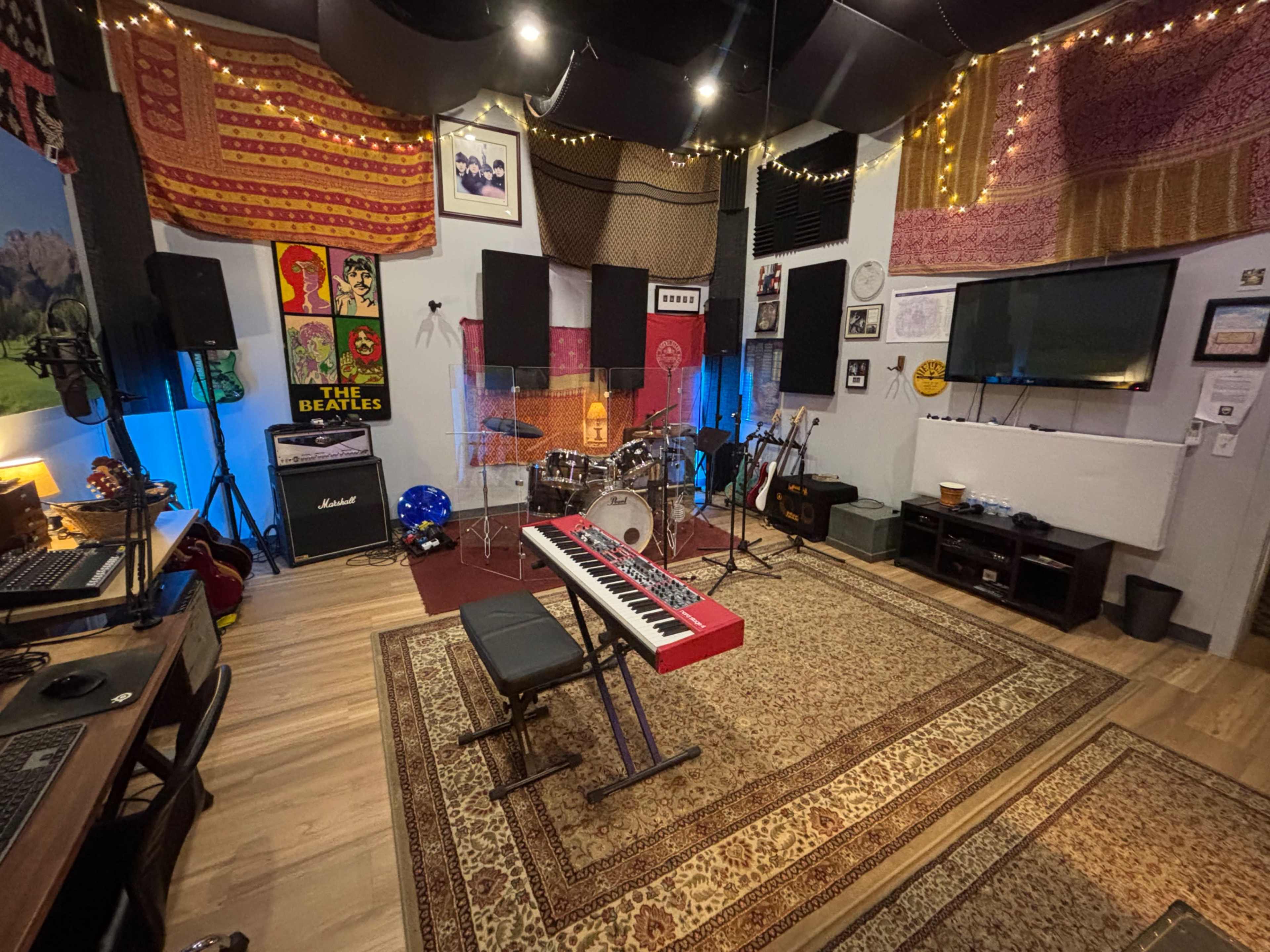 The image shows a music studio featuring a keyboard, drum set, amplifiers, and various sound equipment, decorated with colorful textiles and lighting.