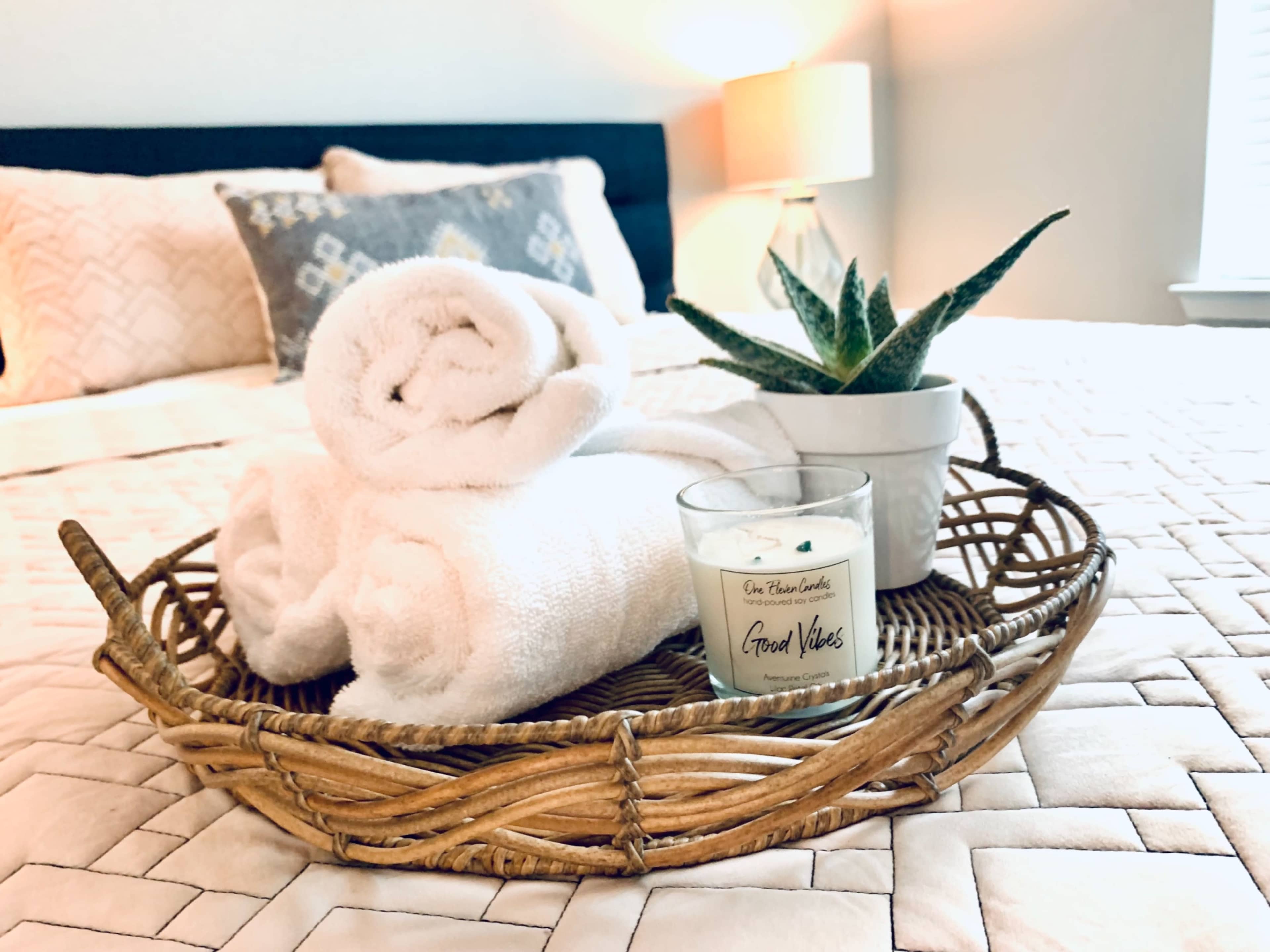 A wicker tray holds rolled white towels, a candle, and a small potted plant on a neatly made bed with a patterned quilt.