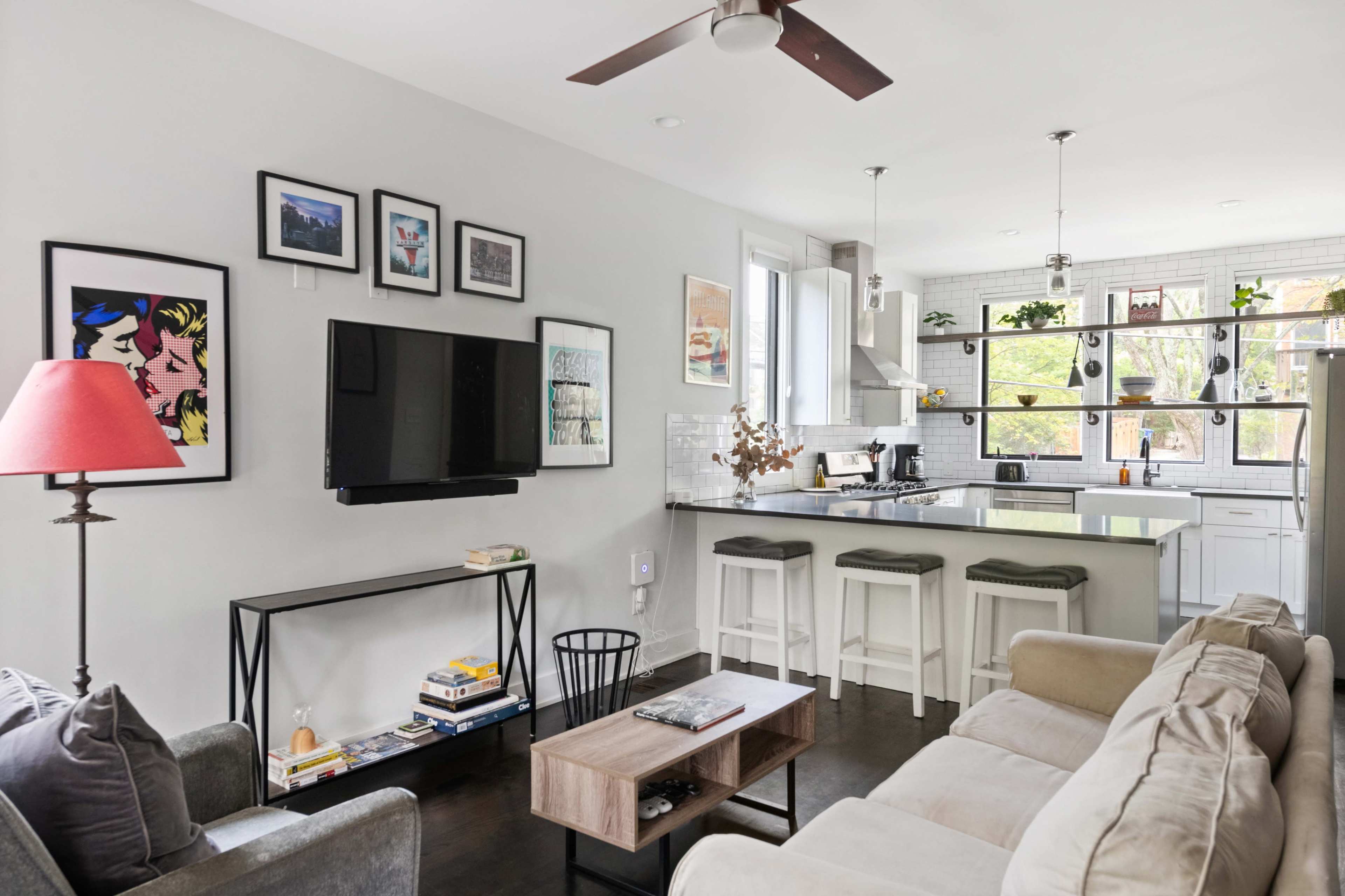 The image shows a modern living room and kitchen area with a mix of artwork on the walls, a television, and bar seating at the kitchen counter.