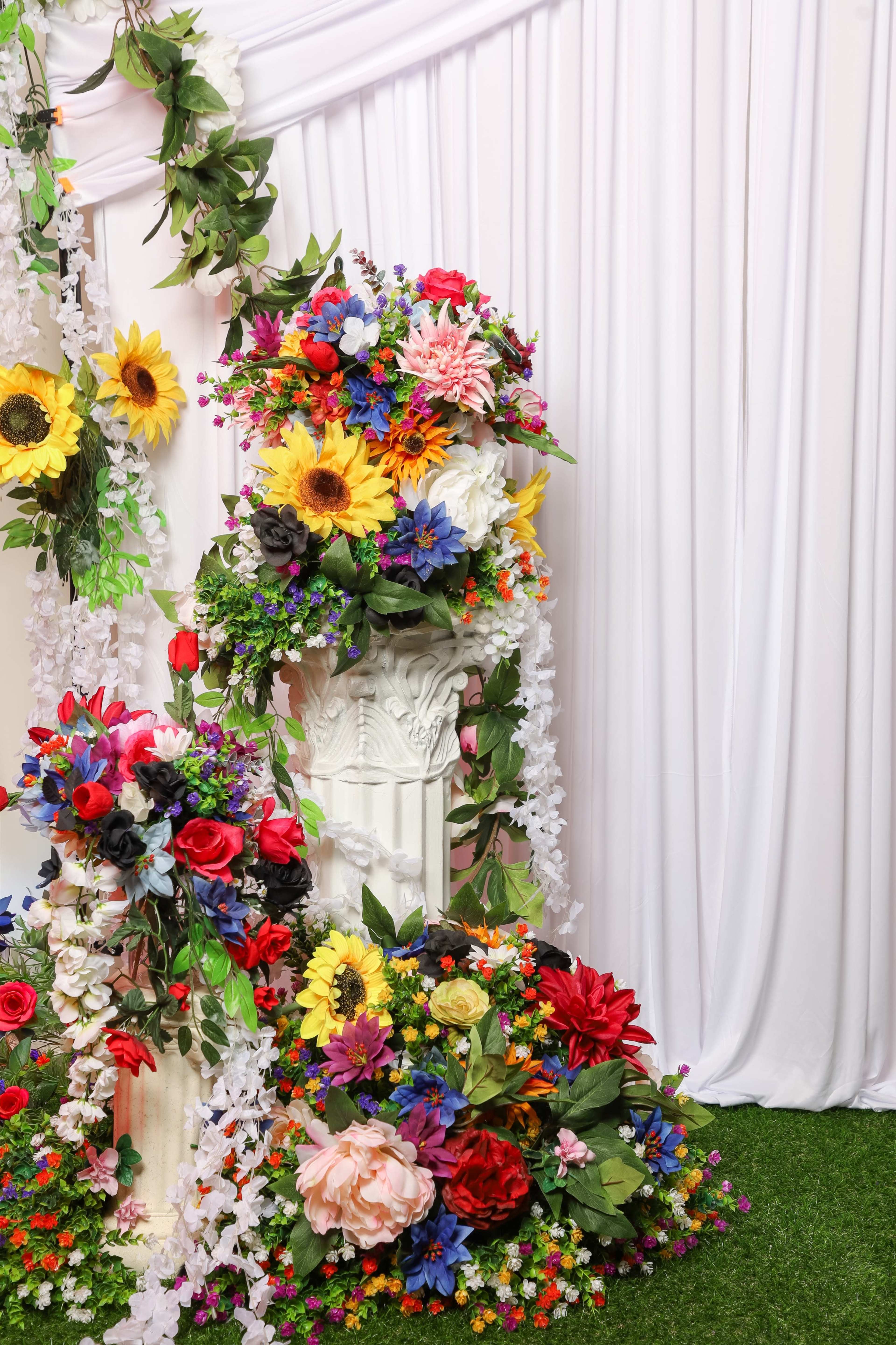 The image shows a decorative floral arrangement featuring various artificial flowers surrounding two ornate white pillars against a white draped backdrop.