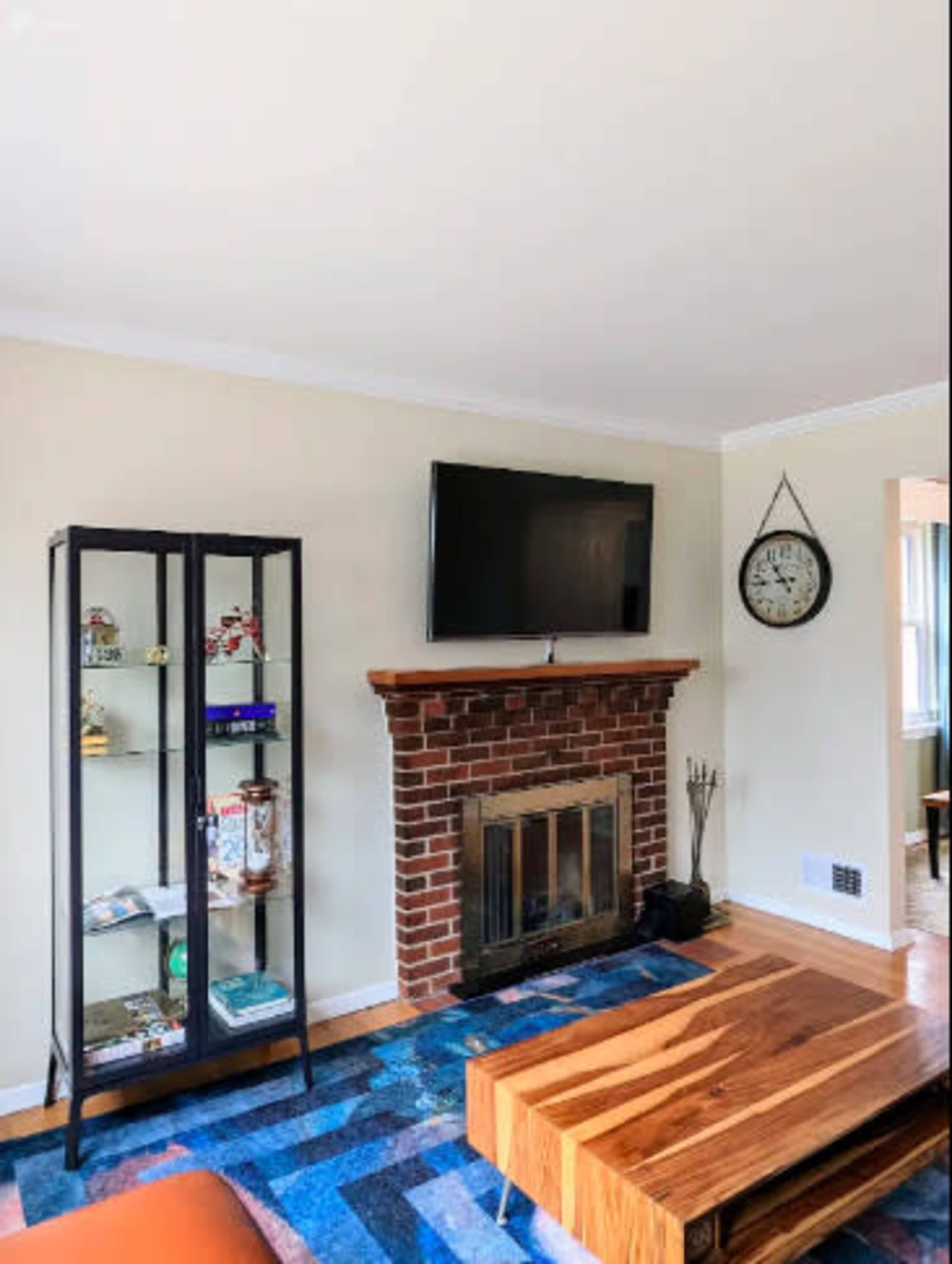 The image shows a living room with a brick fireplace, a mounted television, a glass display cabinet, and a wooden coffee table on a colorful rug.