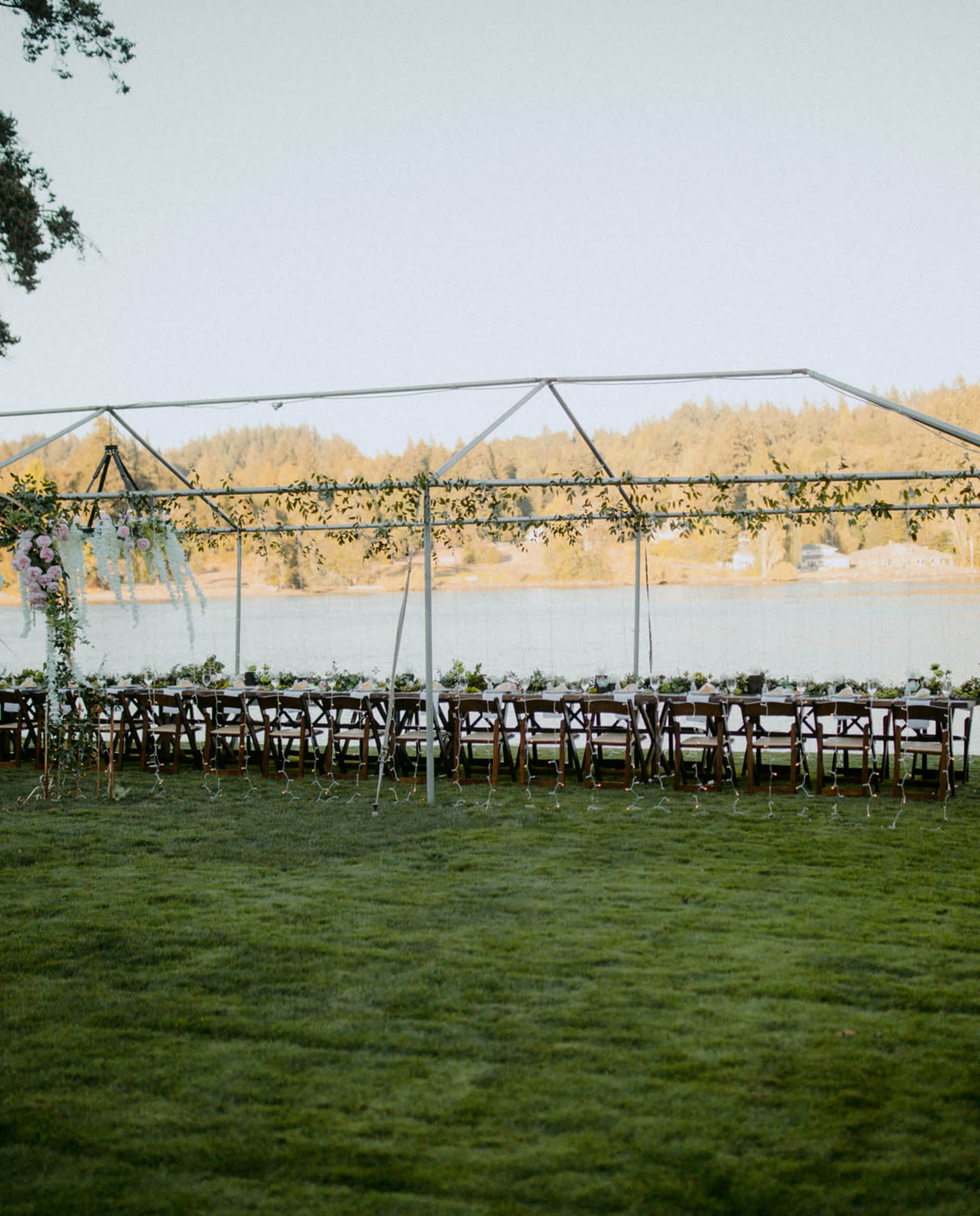 A clear-top tent with wooden chairs is set up on a grassy area overlooking a calm body of water.