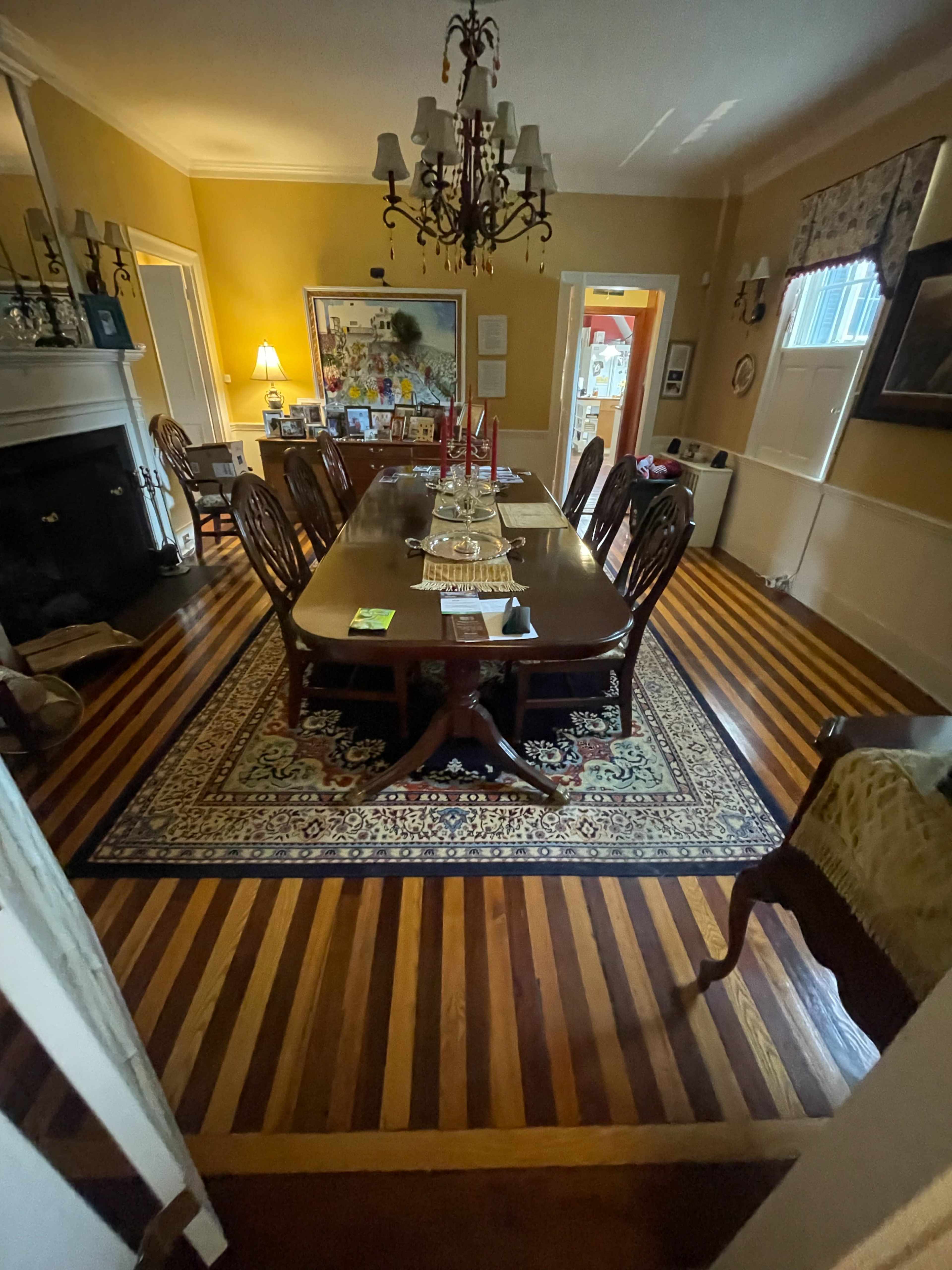 The image shows a dining room with a long wooden table surrounded by chairs, polished hardwood floors, and a large chandelier.