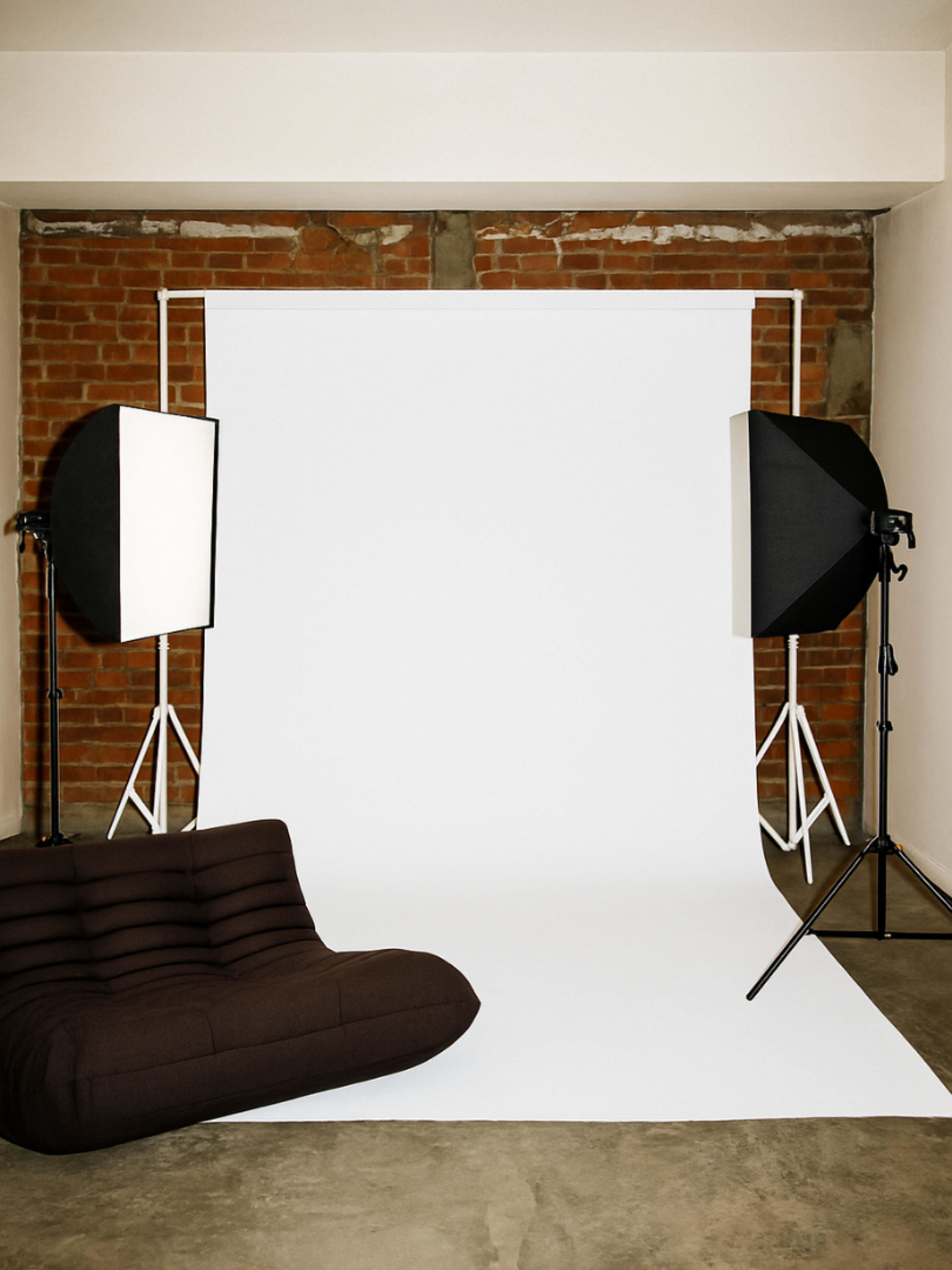 The image shows a photography studio setup with a white backdrop, two light sources on stands, and a brown sofa positioned in front of the backdrop.