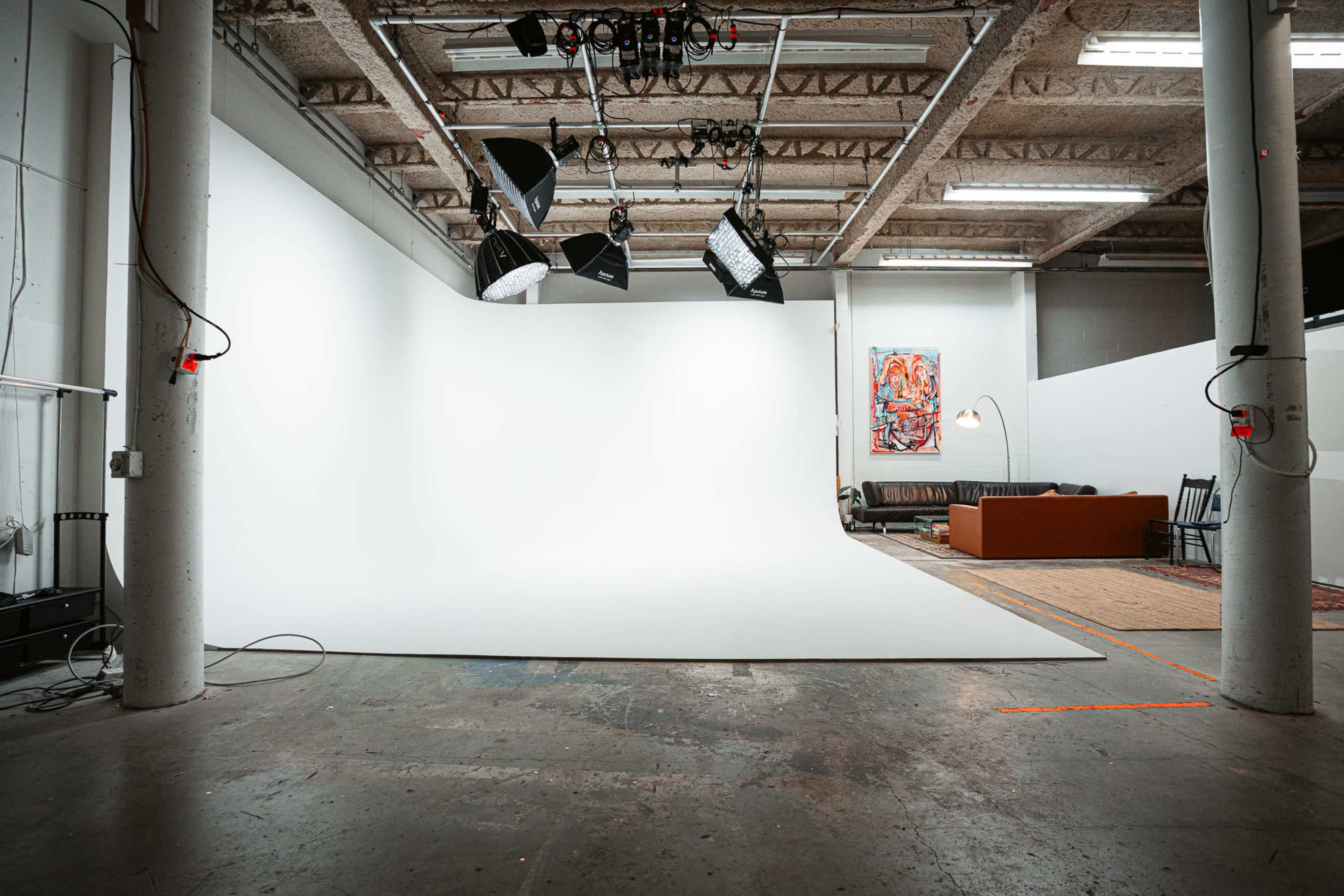 A spacious photography studio features a large white backdrop, bright overhead lights, and a cozy seating area with an orange sofa along one wall.