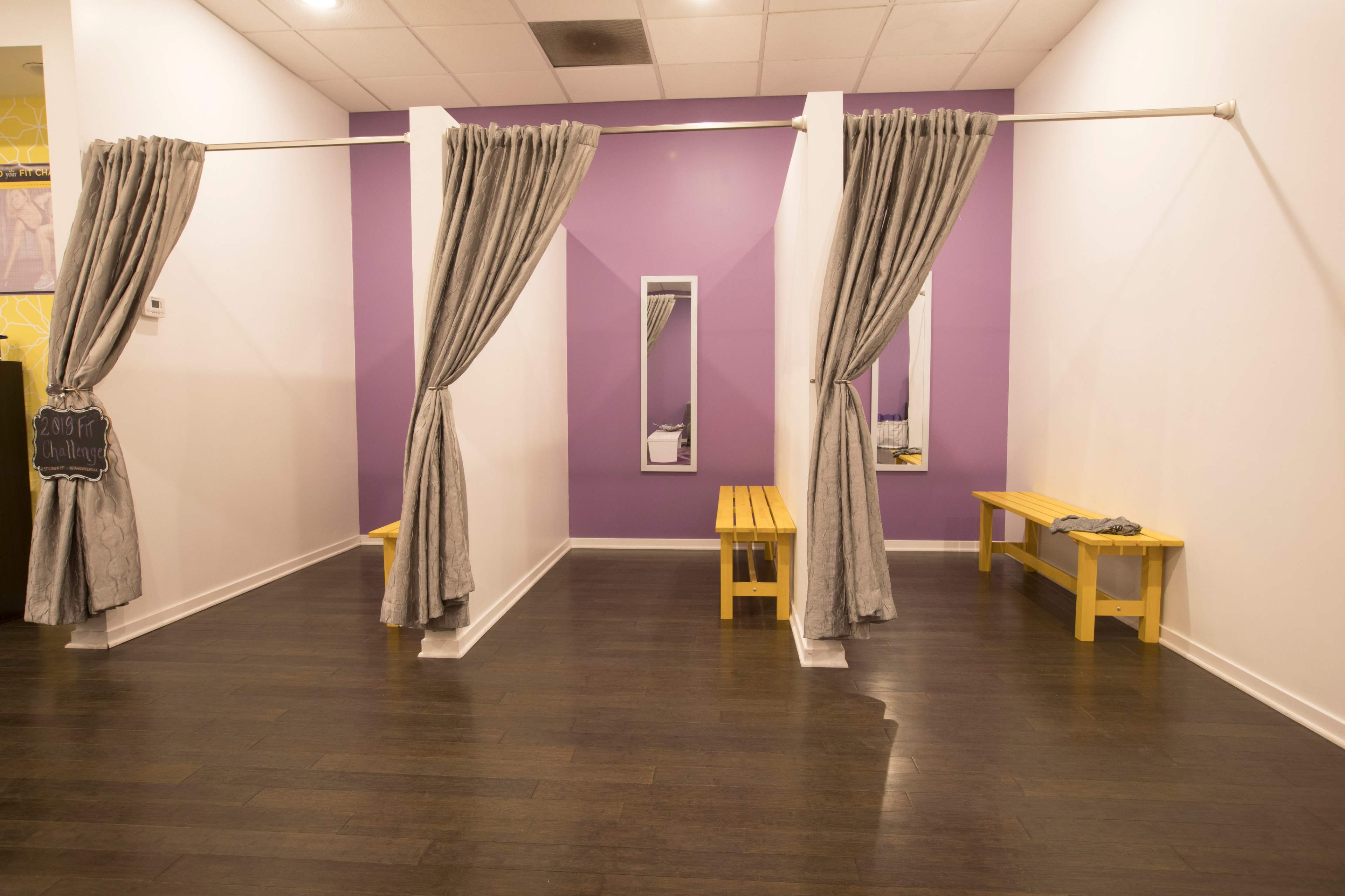A changing room with three curtain-divided sections, each having a wooden bench and a large mirror on the purple wall.
