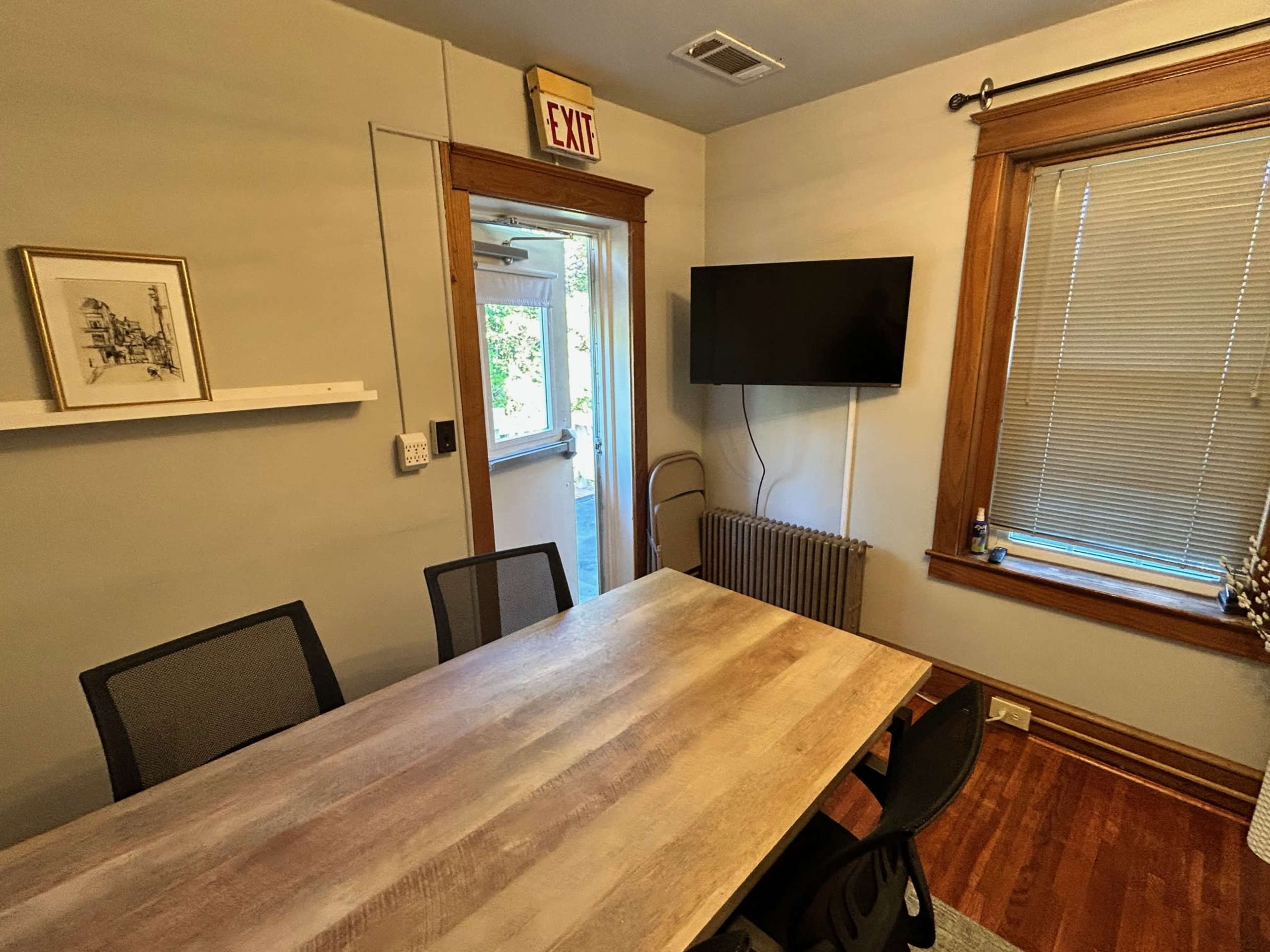 The image shows a small dining or meeting room with a wooden table, black chairs, a wall-mounted TV, and a doorway leading to another space.