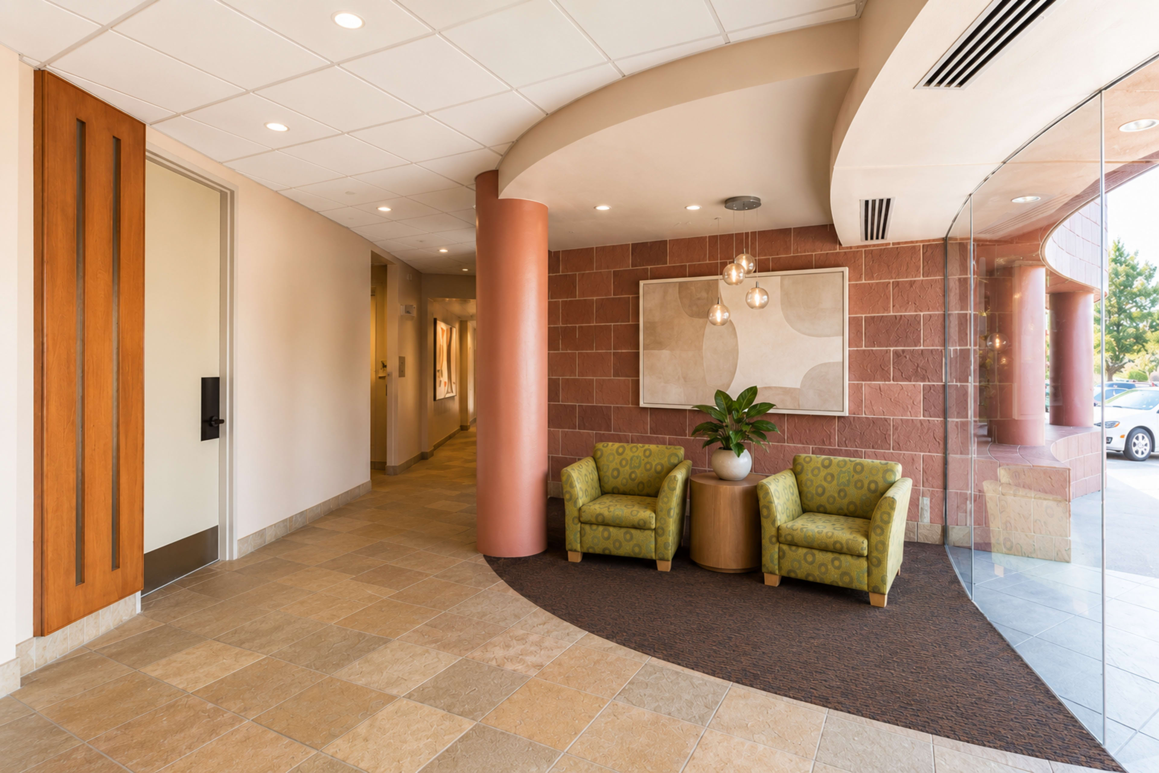 The image shows a modern lobby area with two green patterned chairs, a plant in between them, and a curved wall leading to a corridor.