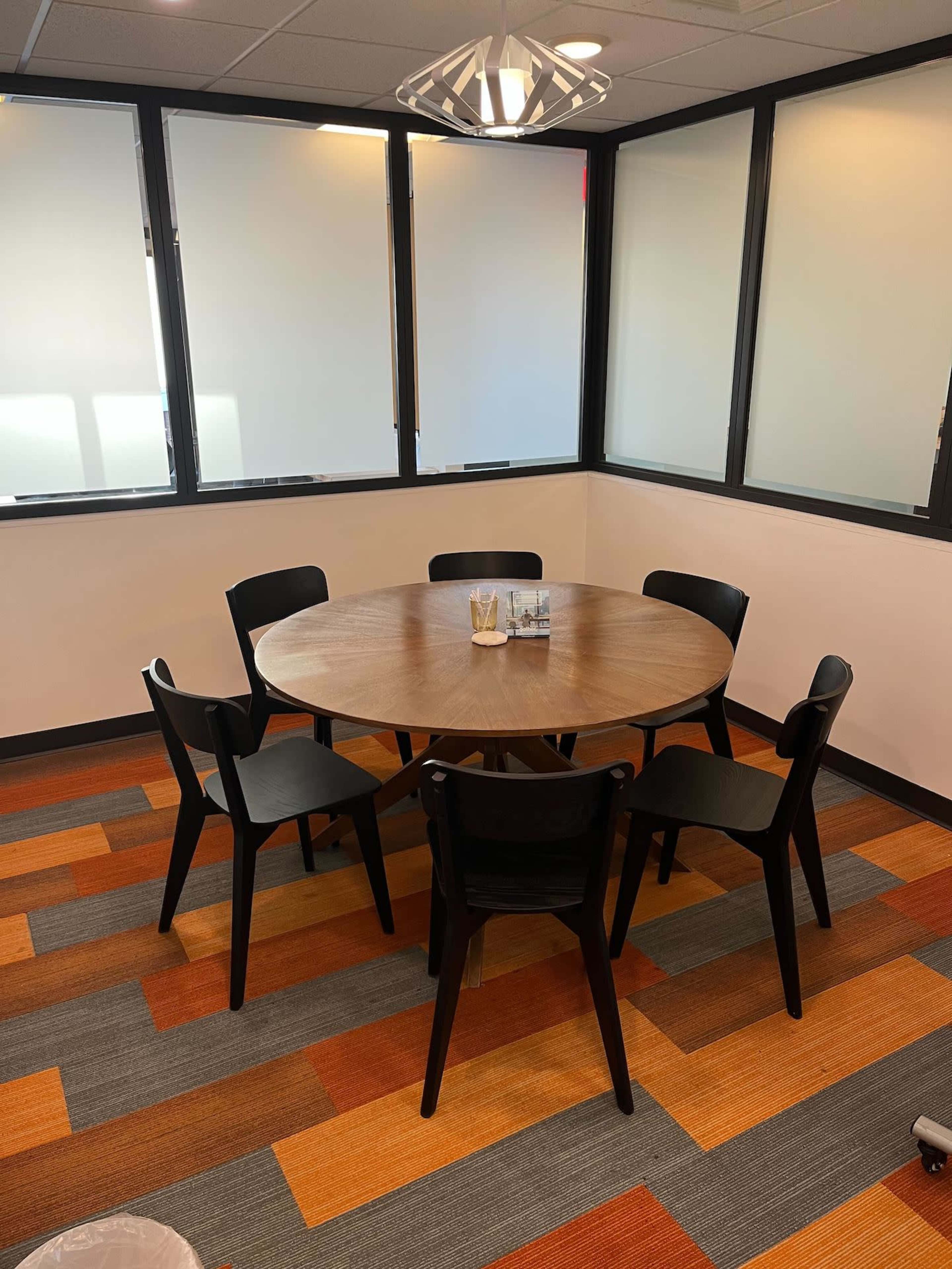 A round wooden table surrounded by six black chairs is positioned in a corner office with large windows.