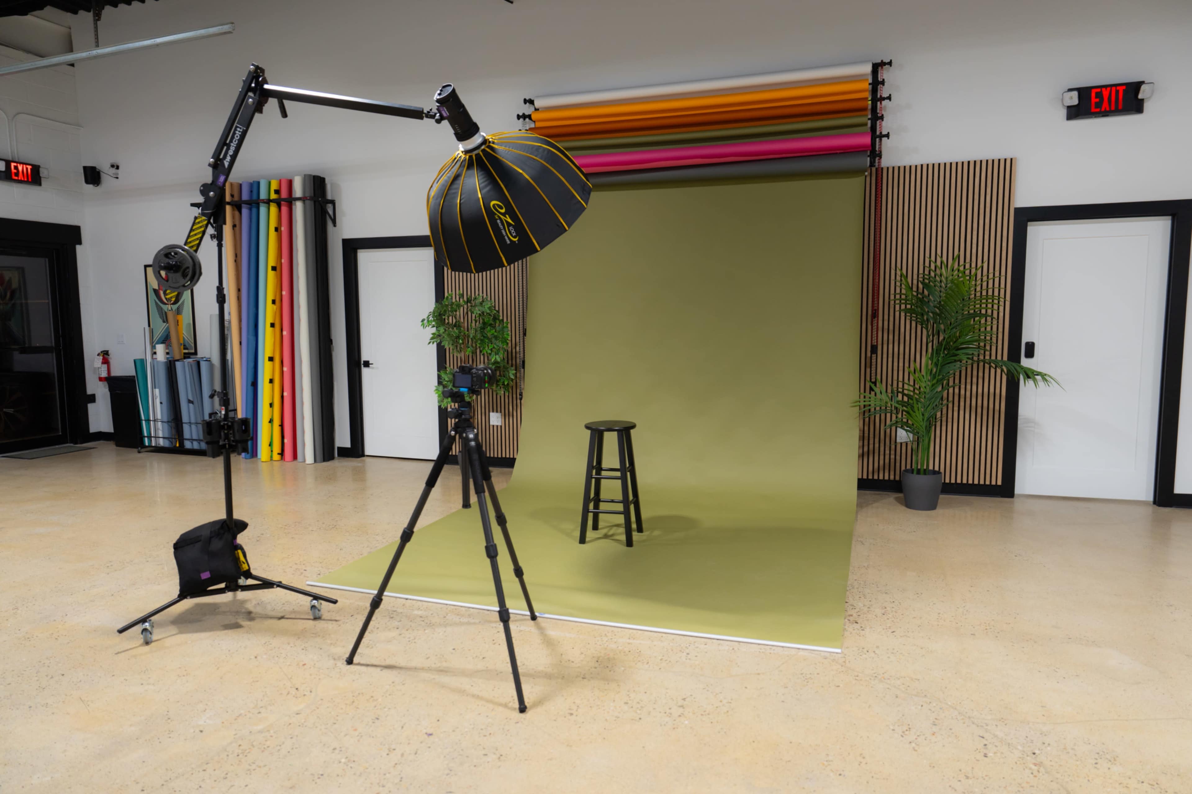 The image shows a photography studio setup with a stool in front of a green backdrop, a camera on a tripod, and a large umbrella light.