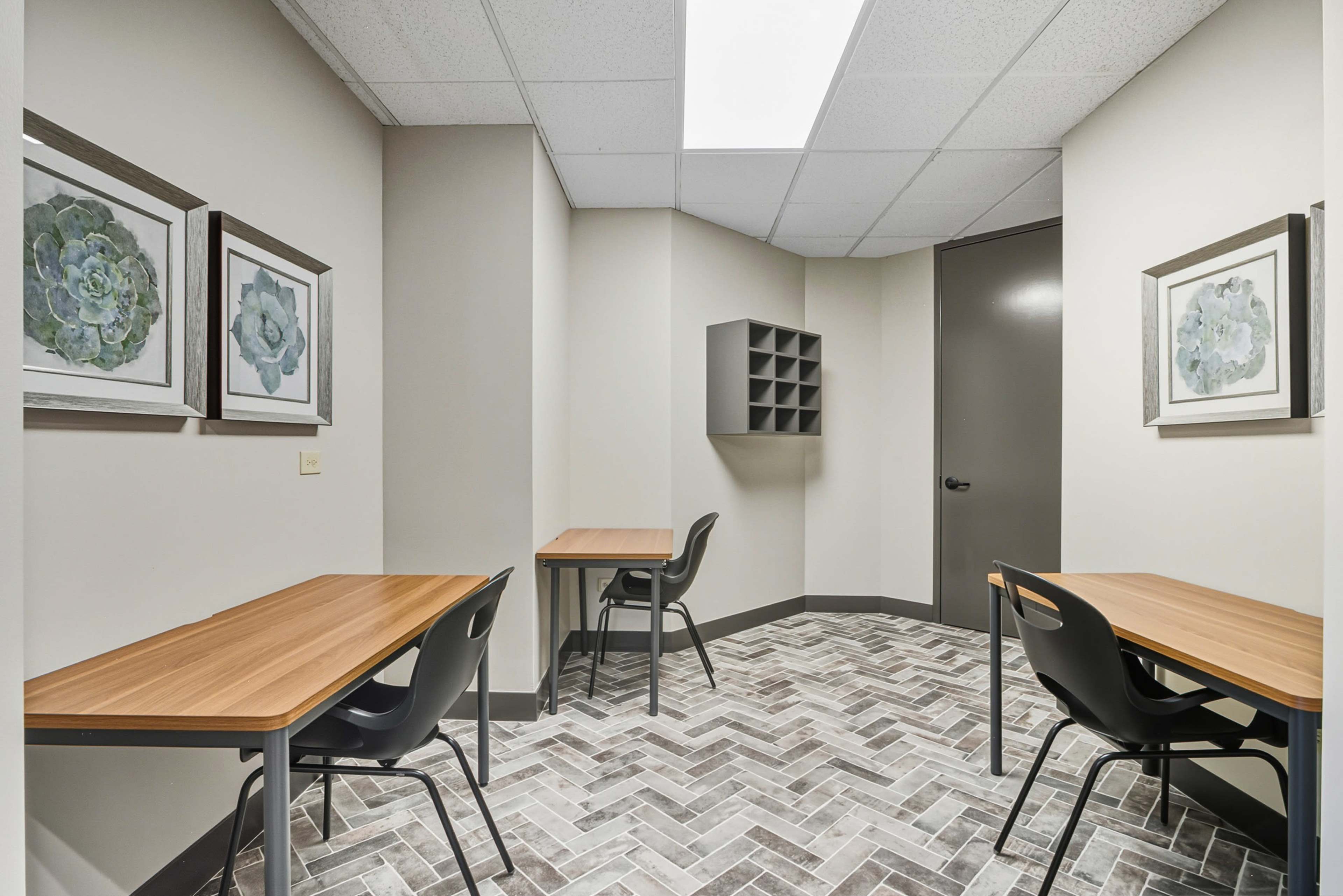 The image shows a small room with two tables and chairs, framed artworks on the walls, and a storage unit by the door.