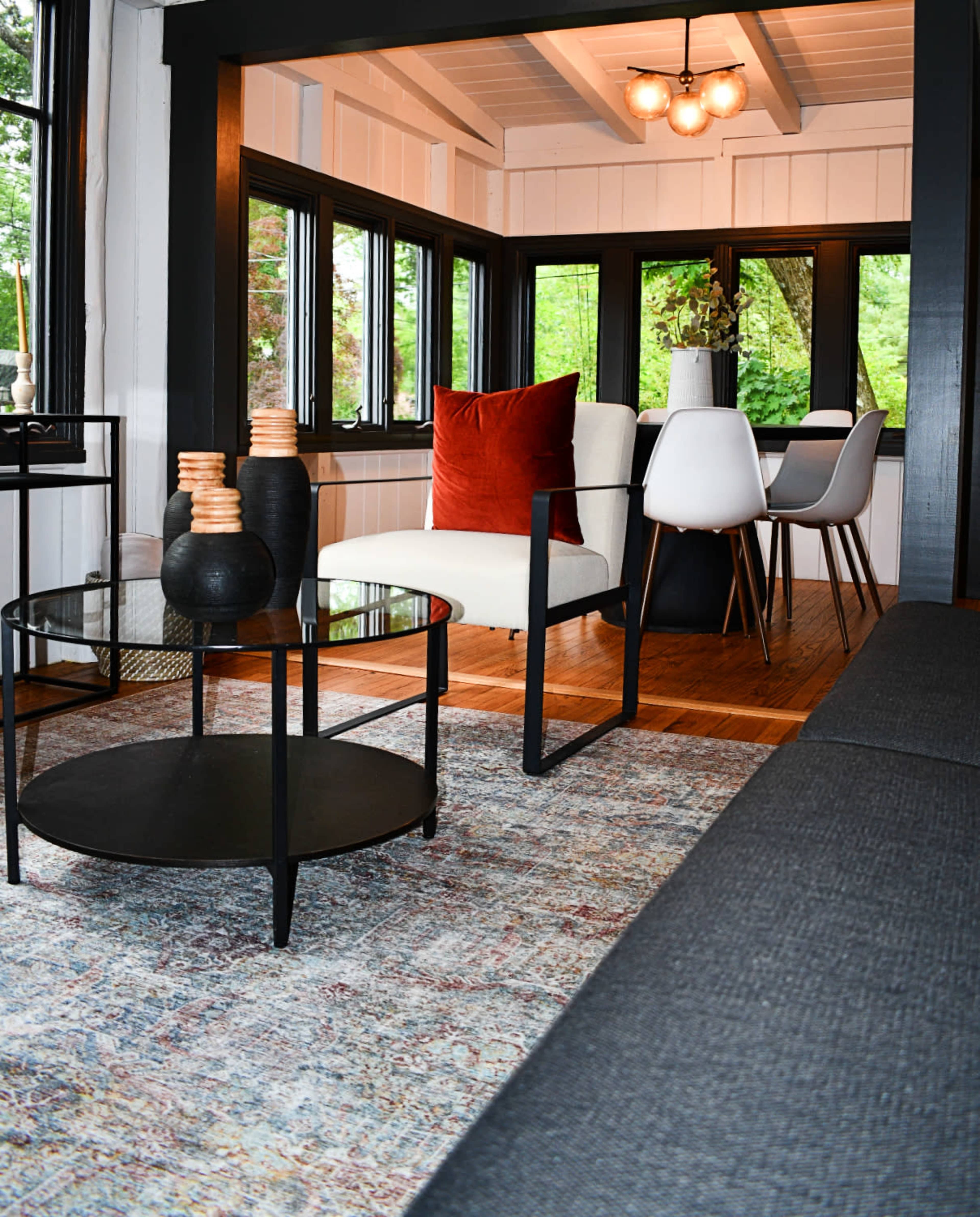 A modern living area features a round coffee table, a white armchair with a red cushion, and a dining table set with white chairs, surrounded by large windows.
