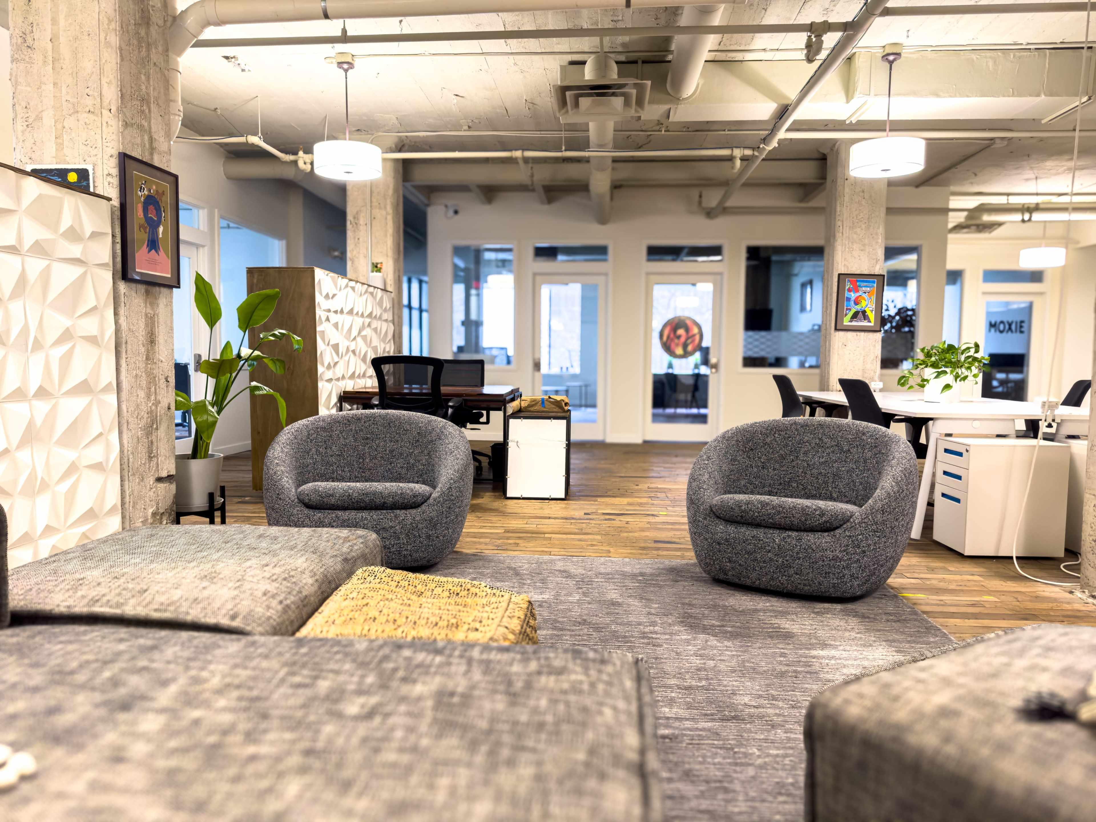 The image shows a modern office space featuring gray upholstered chairs, a wooden floor, and several workstations in the background.