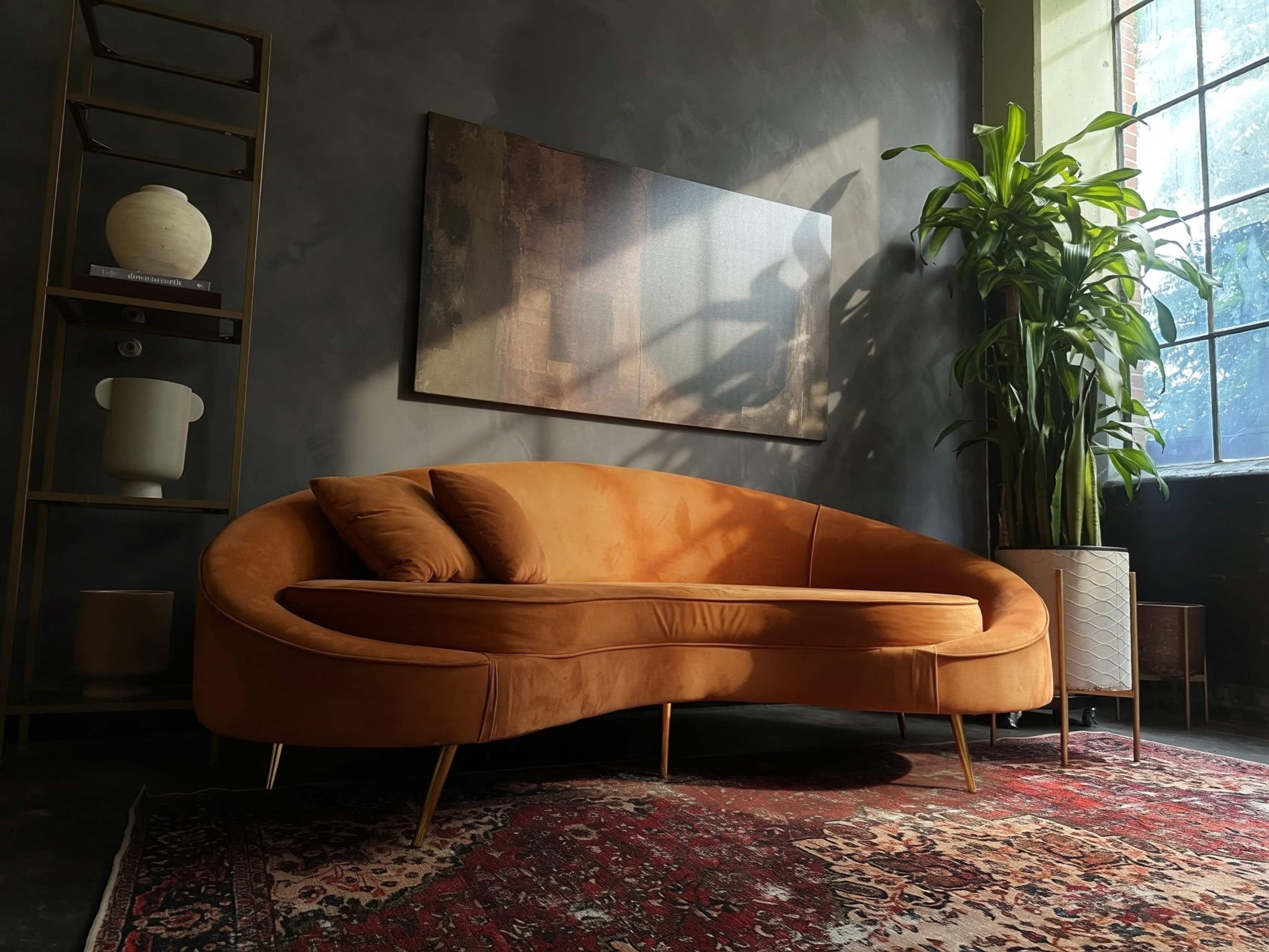 The image features a modern orange sofa positioned in a stylish interior with a large window, a tall plant, a textured wall, and a piece of artwork above the sofa.