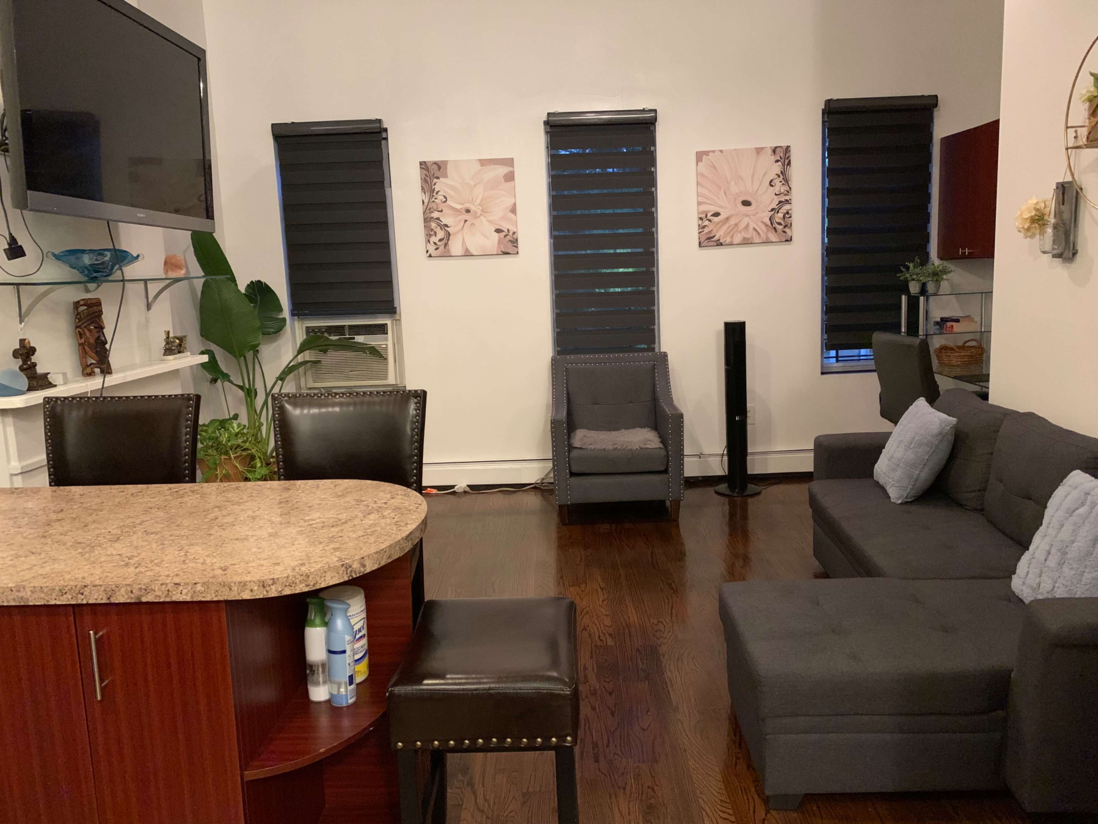 A modern living space with a kitchen bar, two black blinds-covered windows, a gray sectional sofa, and two decorative wall prints.