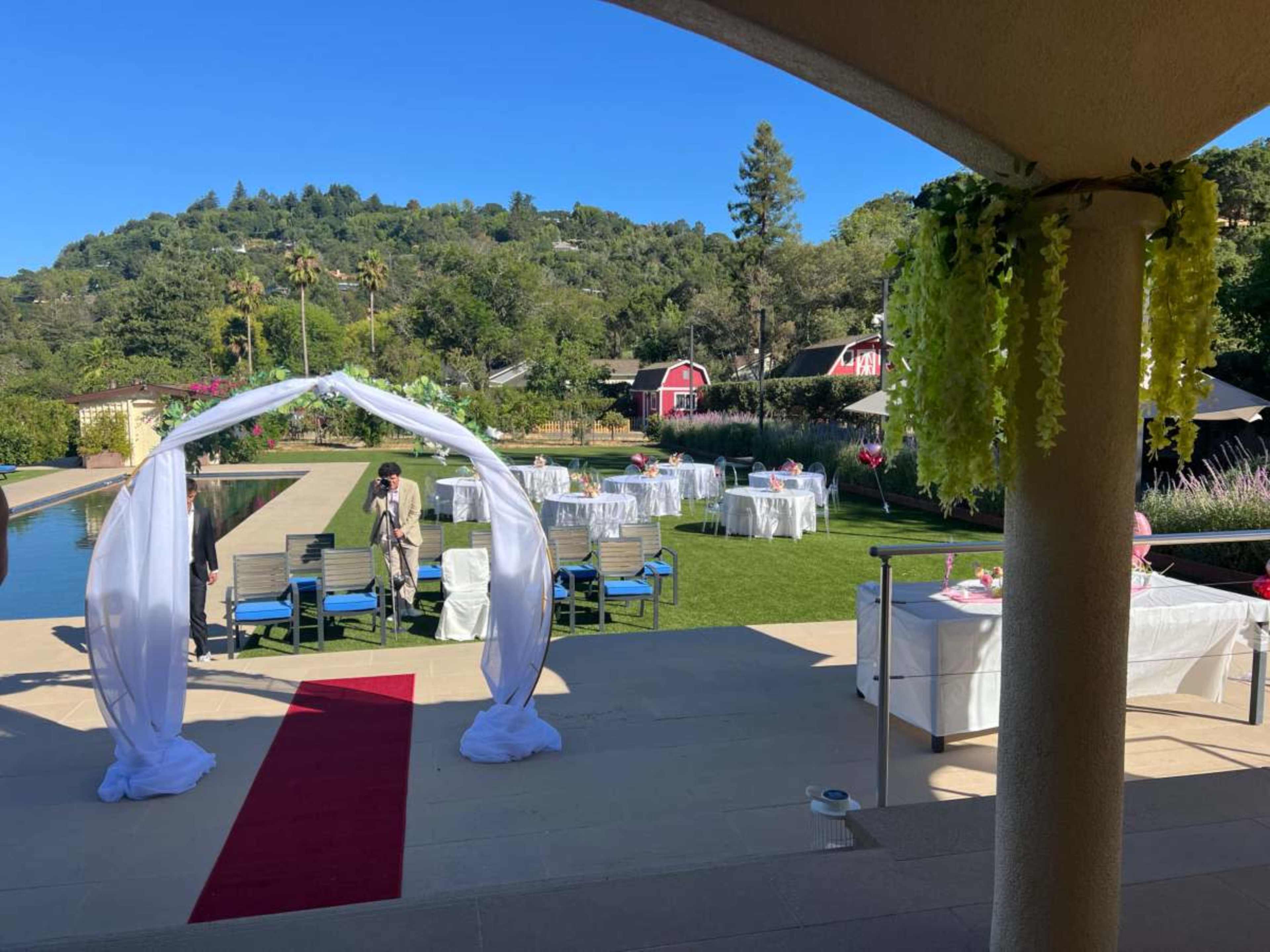 The image shows a decorated outdoor venue with chairs and tables set up on a lawn, a white floral arch near a pool, and mountains in the background.