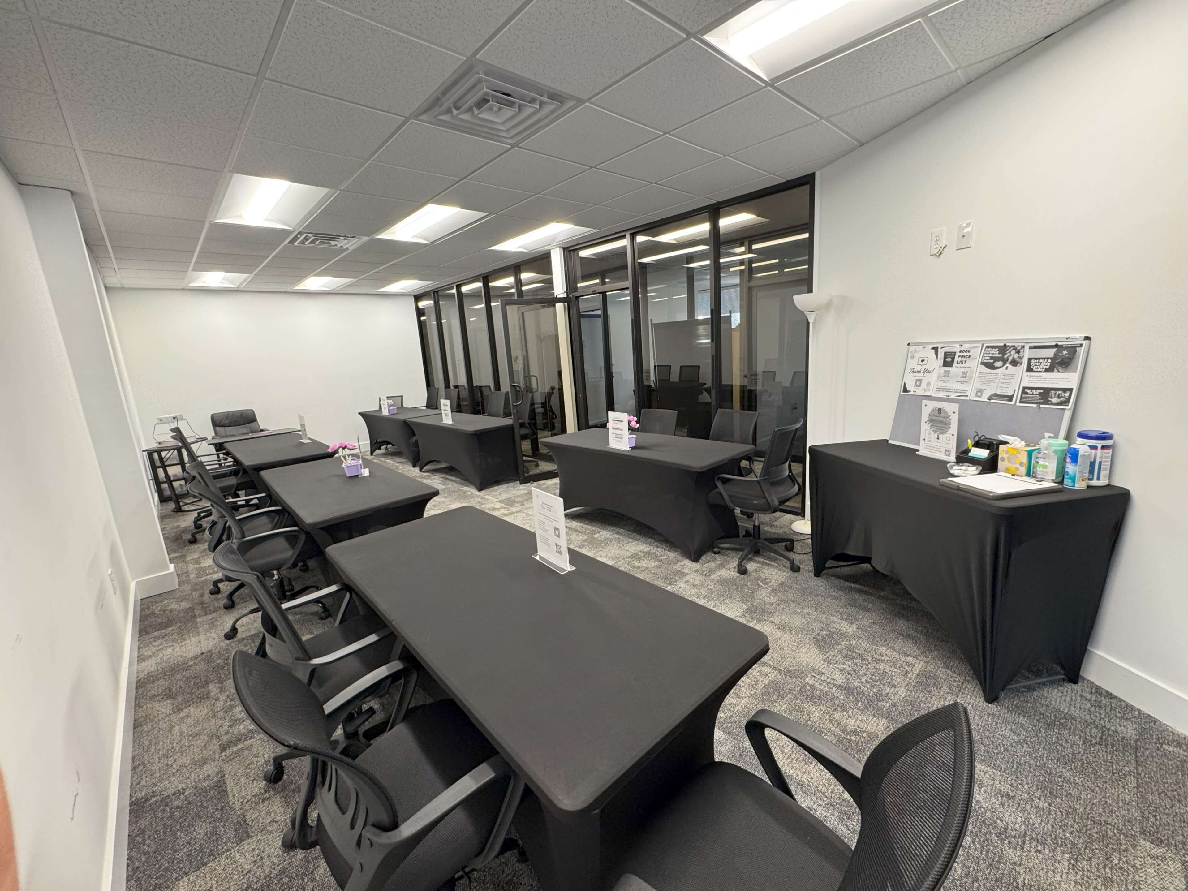 The image shows a modern office space with several black-covered tables arranged in neat rows, surrounded by gray carpeting and large windows.