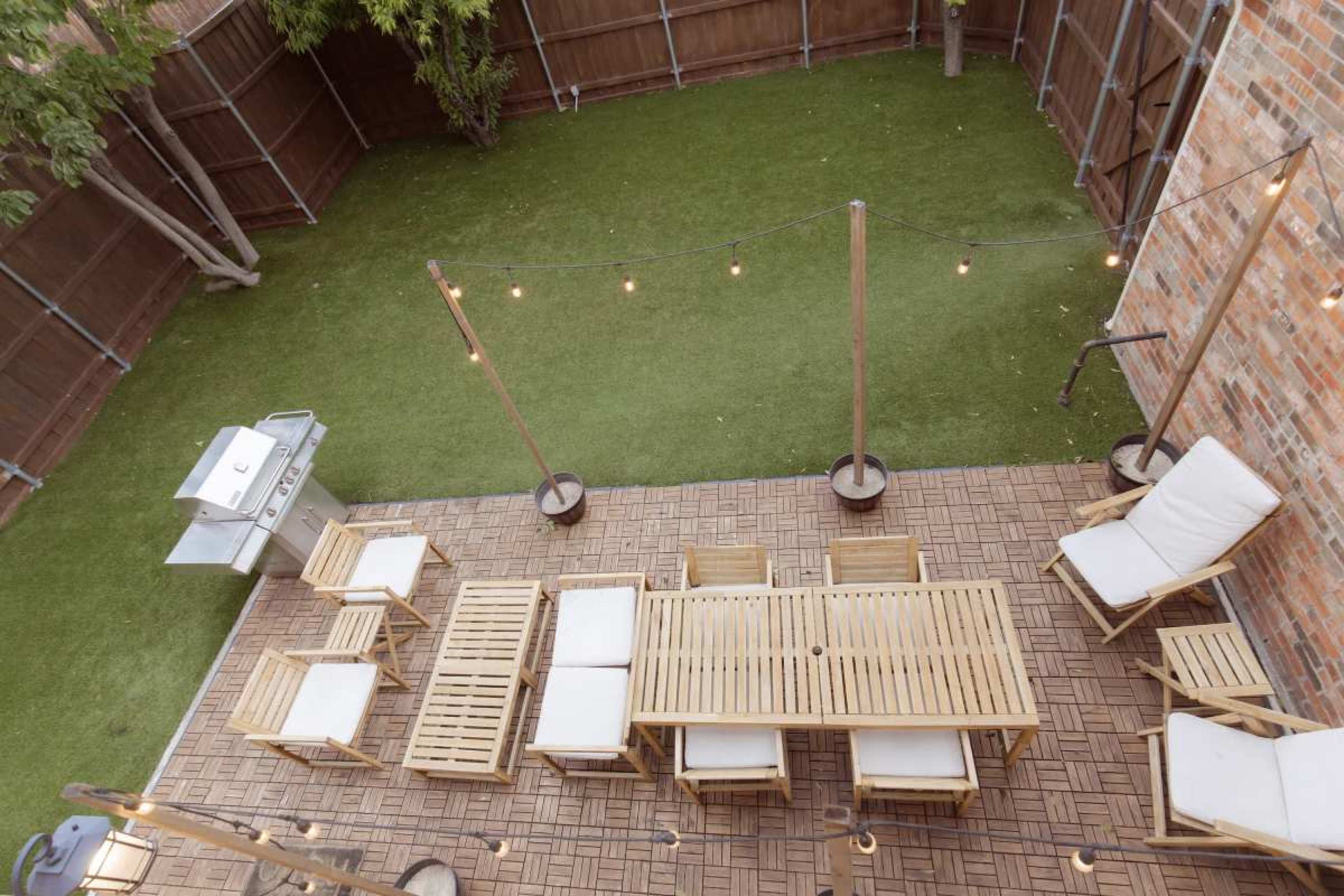The image shows a backyard patio with wooden furniture, string lights, and a grill, surrounded by green grass and wooden fencing.