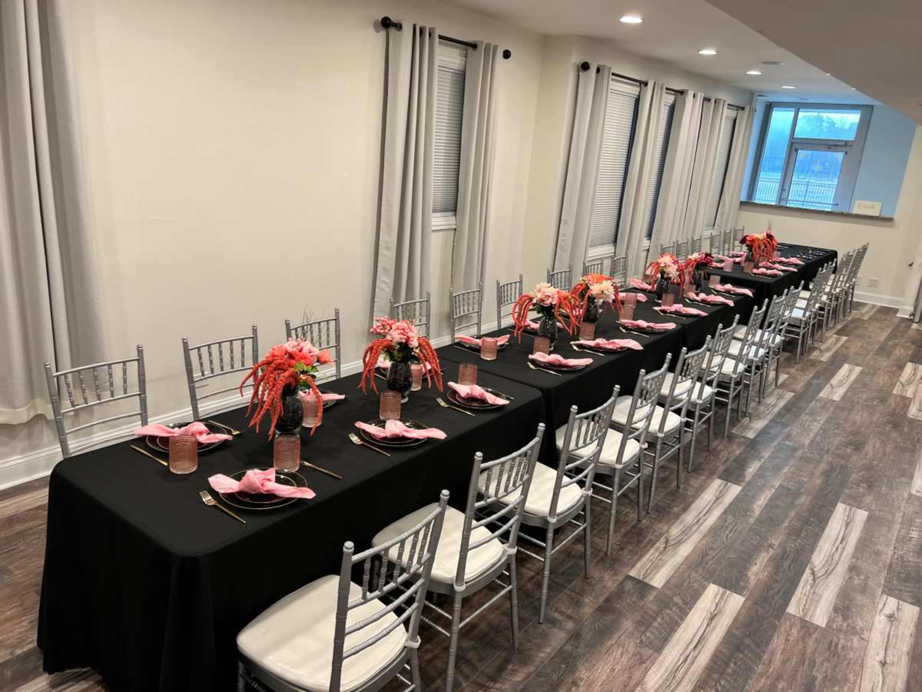 A long, rectangular dining table is set with black tablecloths, pink napkins, and flower centerpieces, surrounded by silver chiavari chairs in a well-lit room.