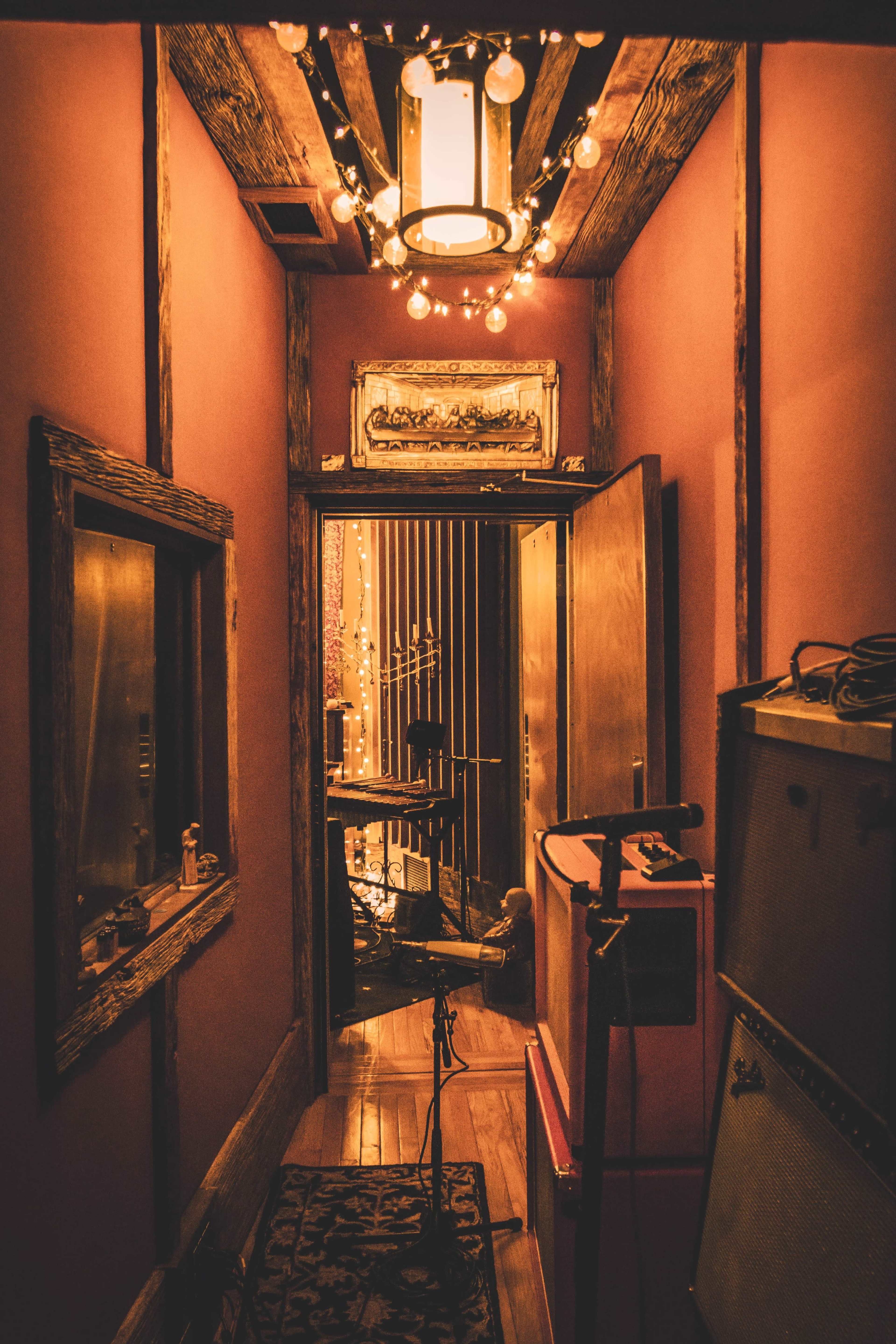 The image shows a dimly lit hallway leading to a recording studio, with wooden beams, a decorative light fixture, and musical equipment positioned along the walls.