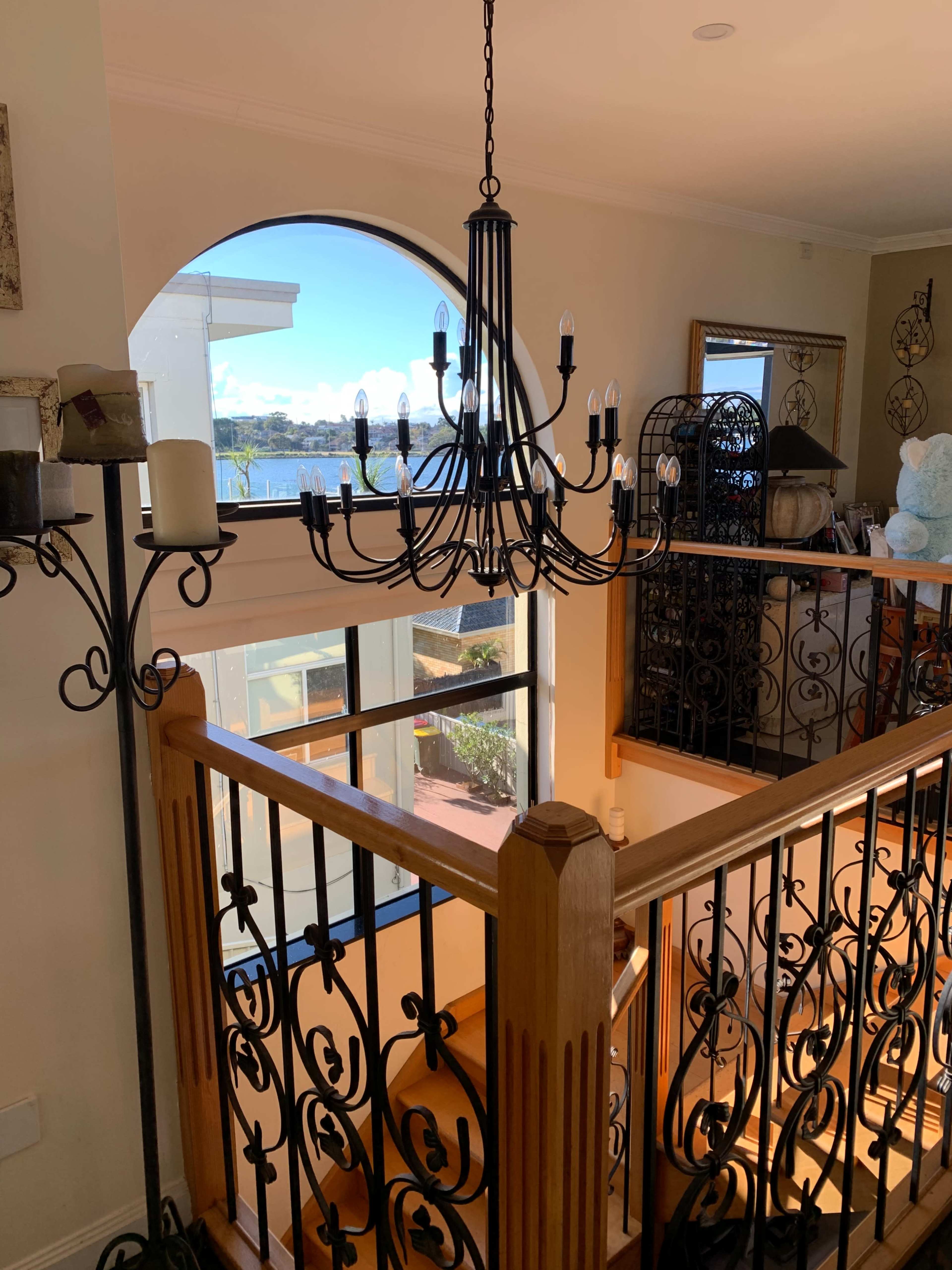 A chandelier hangs above a staircase with wrought iron railings, leading to a window that overlooks a waterfront view.