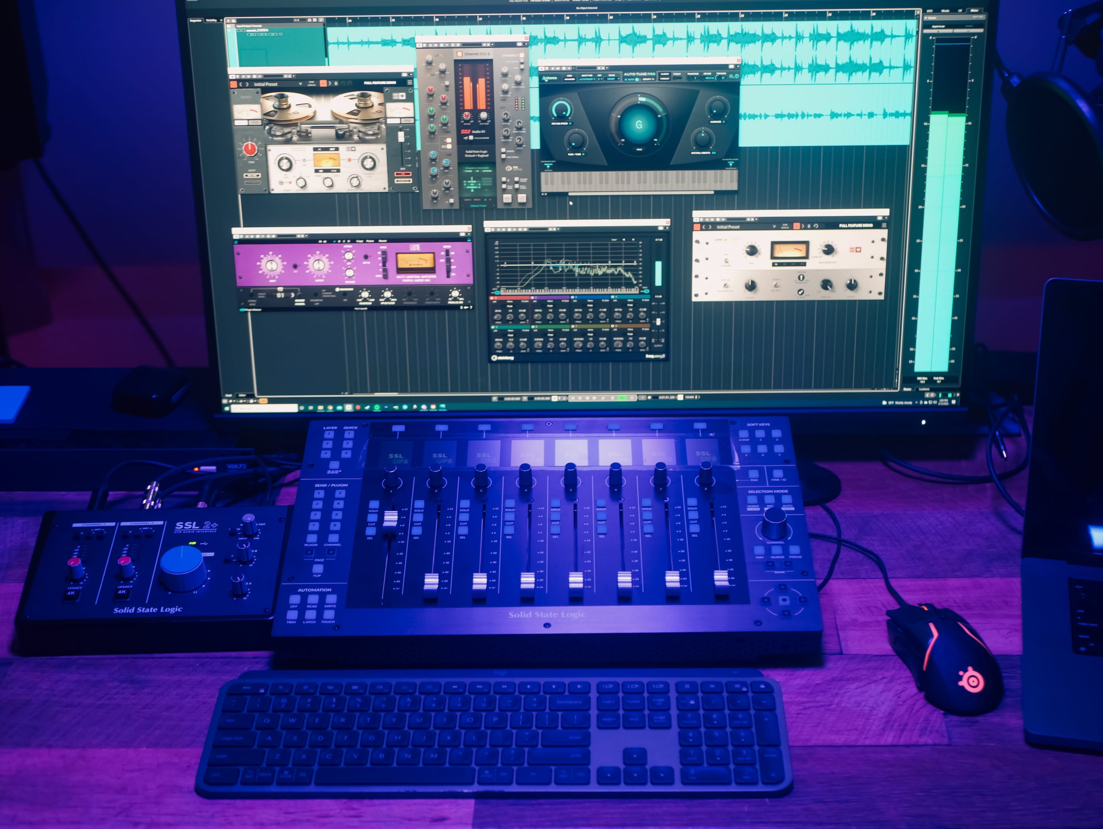 A computer setup with a large monitor displaying audio editing software and a digital audio workstation controller on a wooden desk.