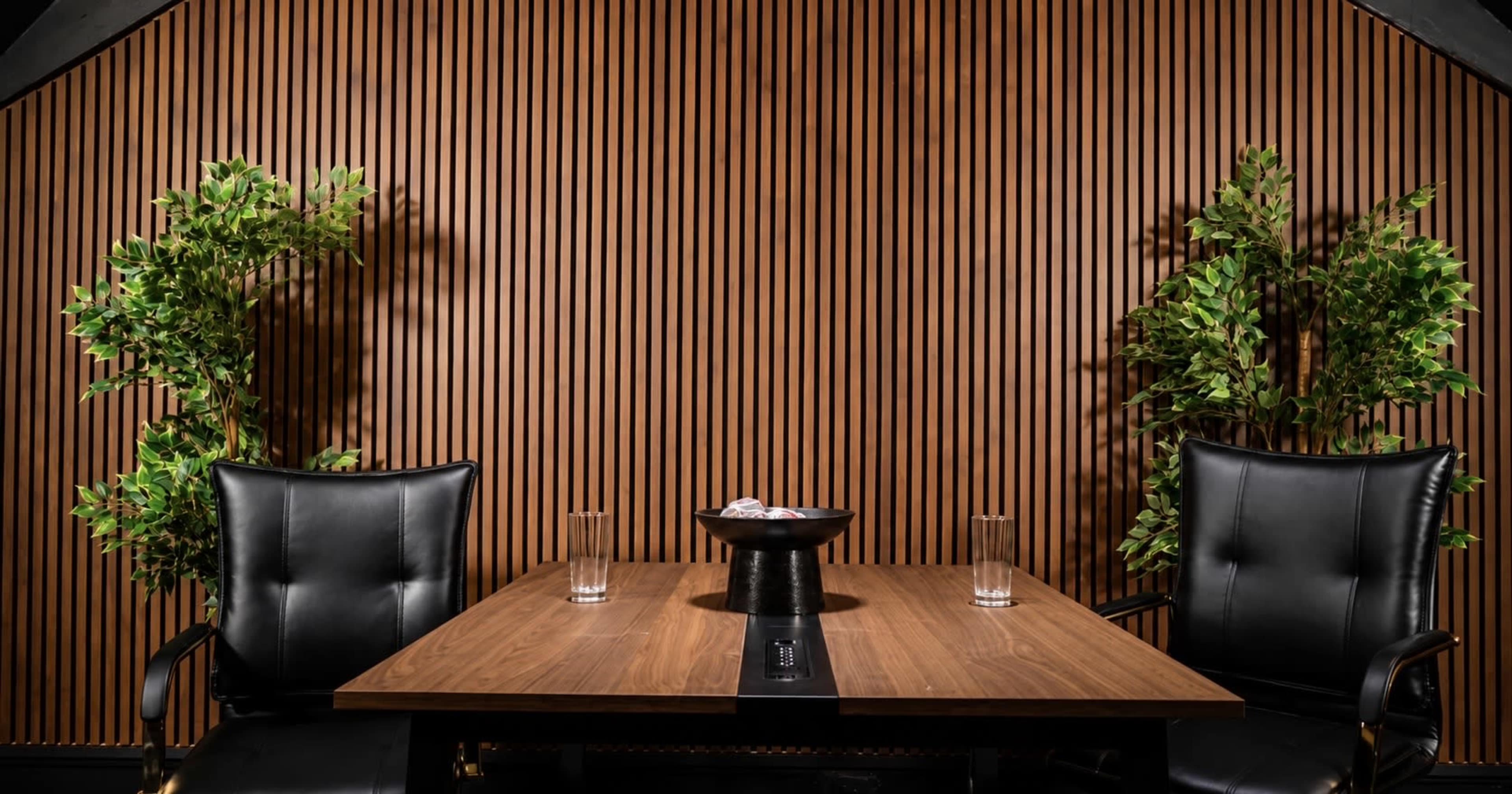 A wooden table with two black leather chairs is set against a slatted wooden wall, accompanied by two potted plants on either side.