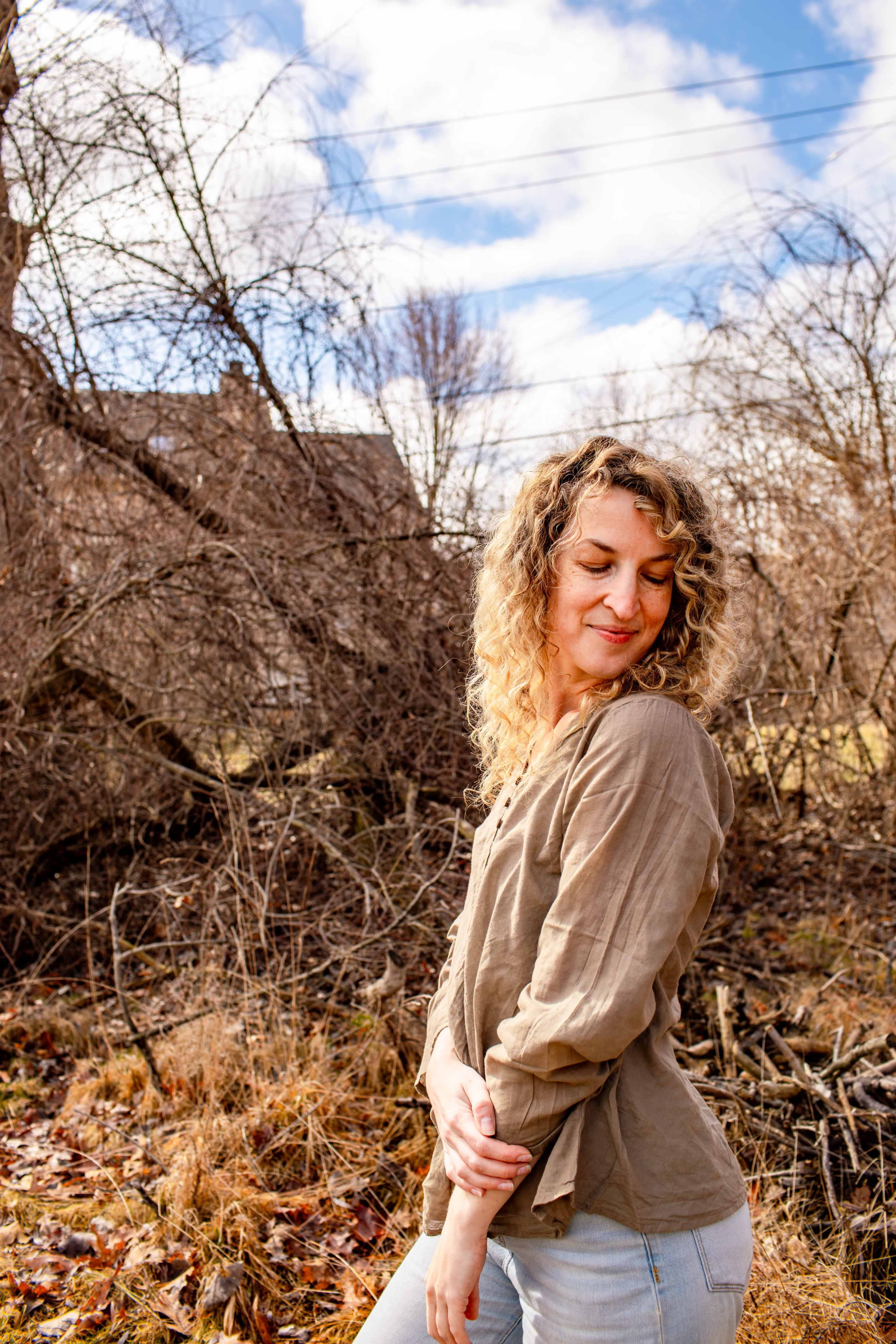 A woman with curly hair stands outdoors in front of a tangle of trees and underbrush, wearing a light brown top and light blue jeans.