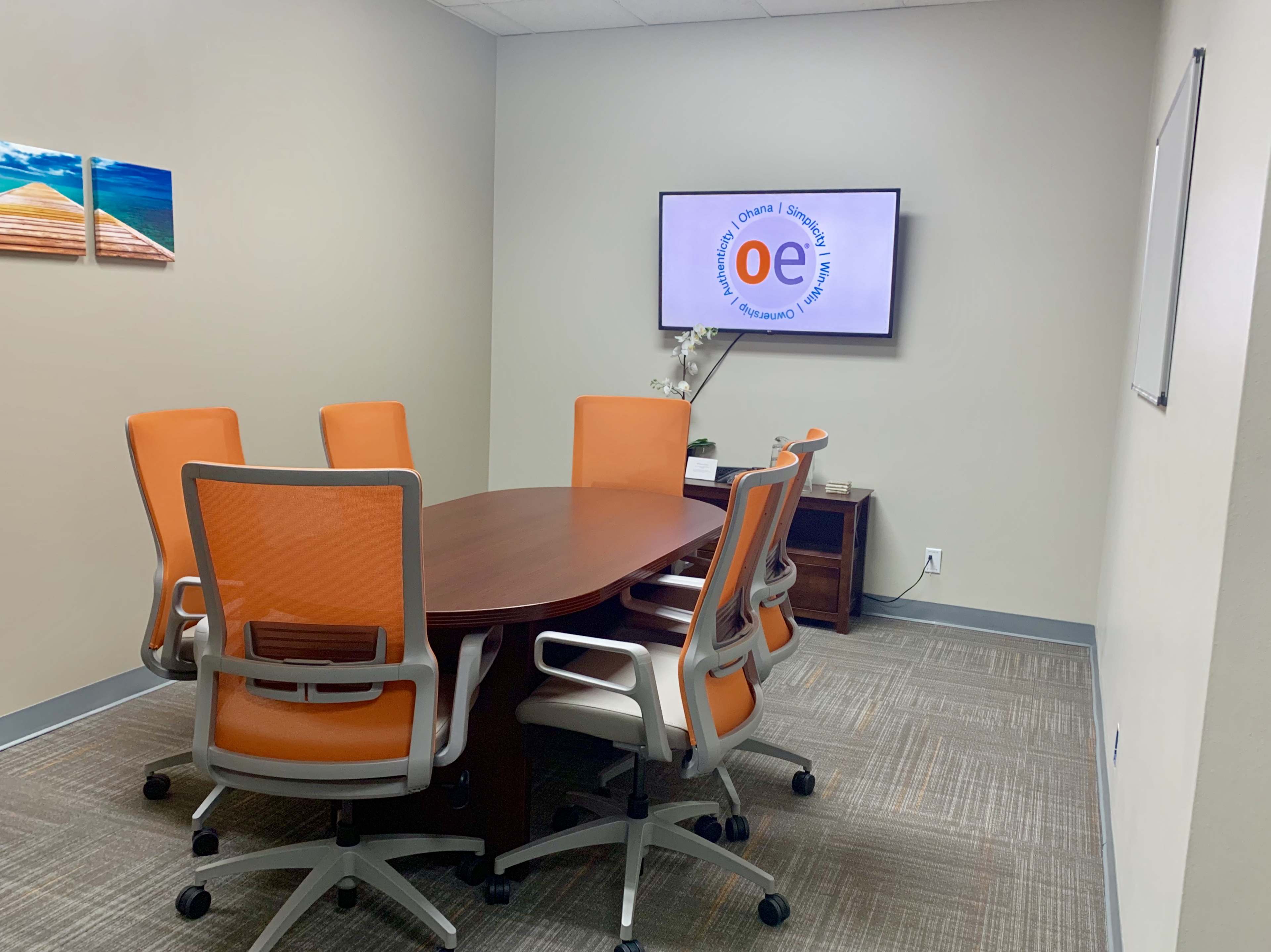 A conference room is depicted with a round wooden table surrounded by six orange chairs, a television displaying a logo, and a whiteboard on the wall.