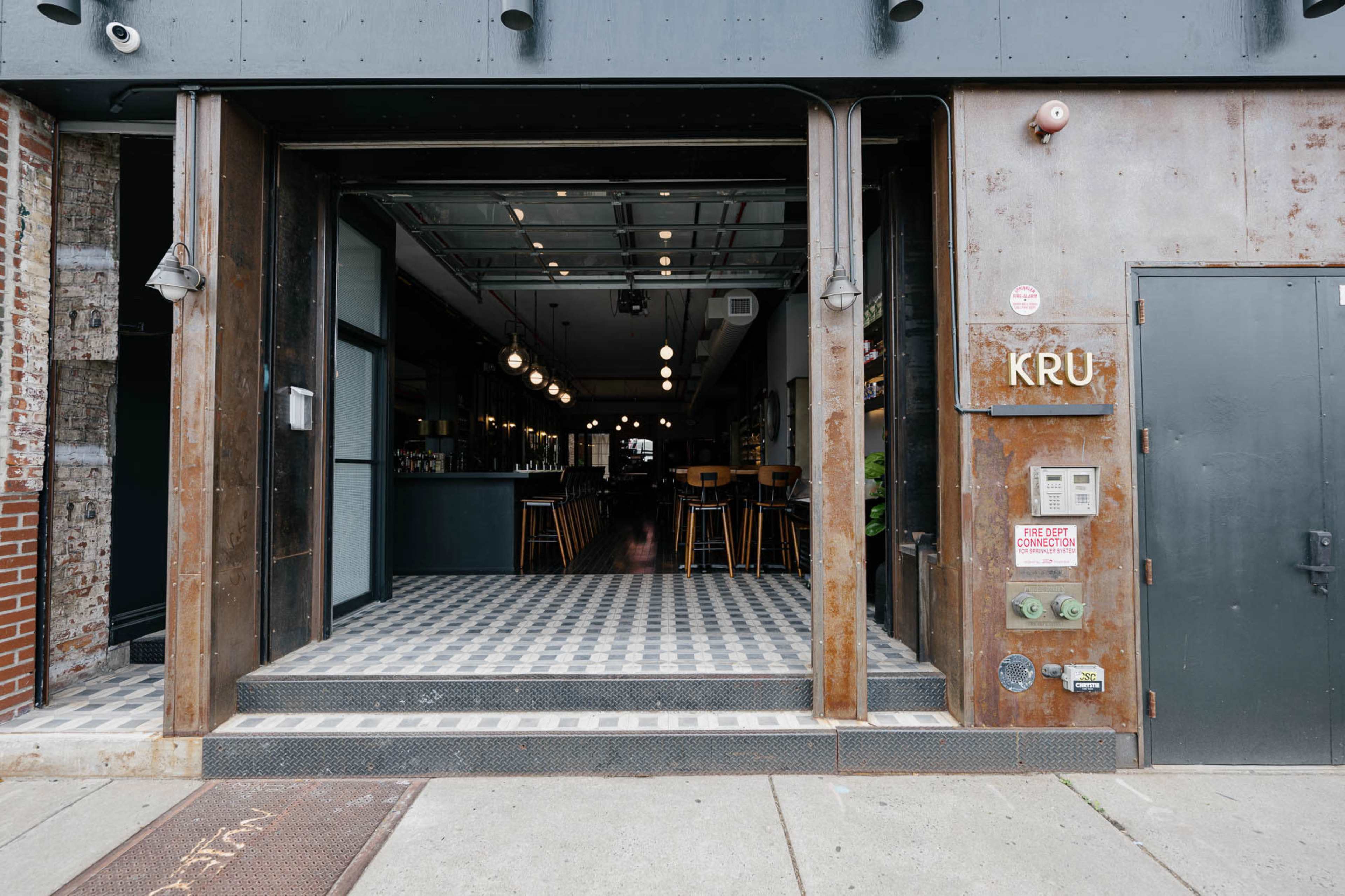 The image shows the entrance of a restaurant called "KRU," featuring large open doors that reveal a modern interior with a checkered floor and bar seating.