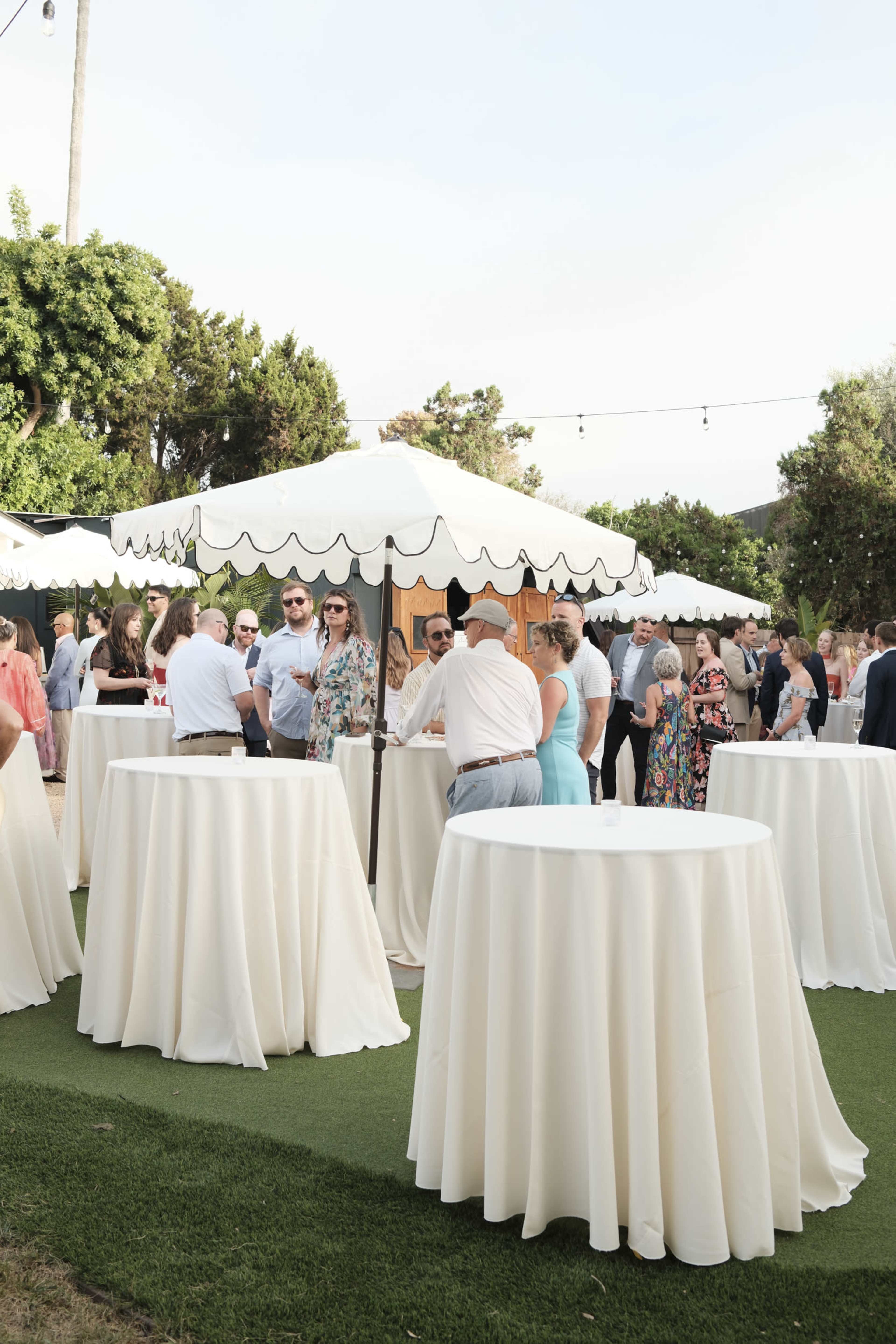 A crowd of people socializes around several high-top tables under white canopies at an outdoor event.